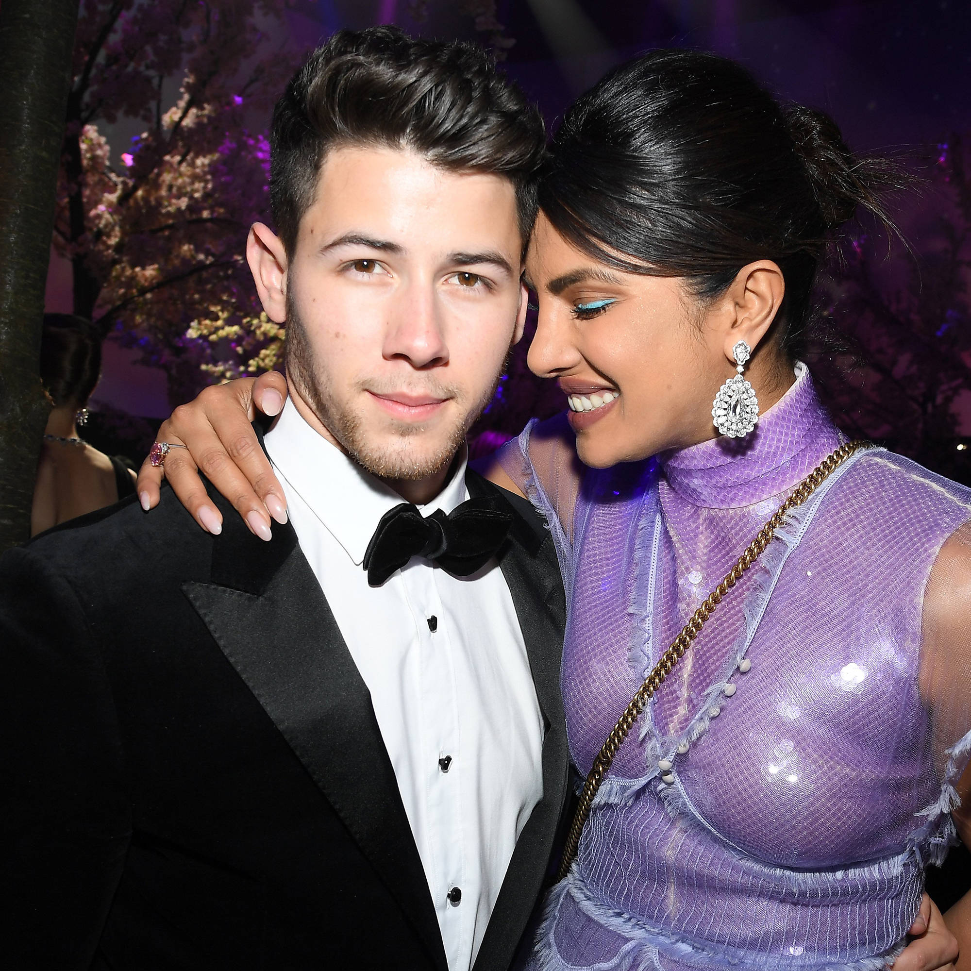 Priyanka 桌布 壁紙 — 一對魅力情侶在夜間活動中緊密擺姿勢，背景有紫色燈光和櫻花。