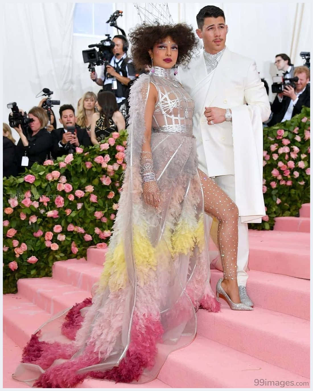 Met Gala 桌布 壁紙 — 一對魅力情侶在粉紅色地毯樓梯上擺姿勢，兩旁佈滿粉紅玫瑰，出席高級時裝活動。