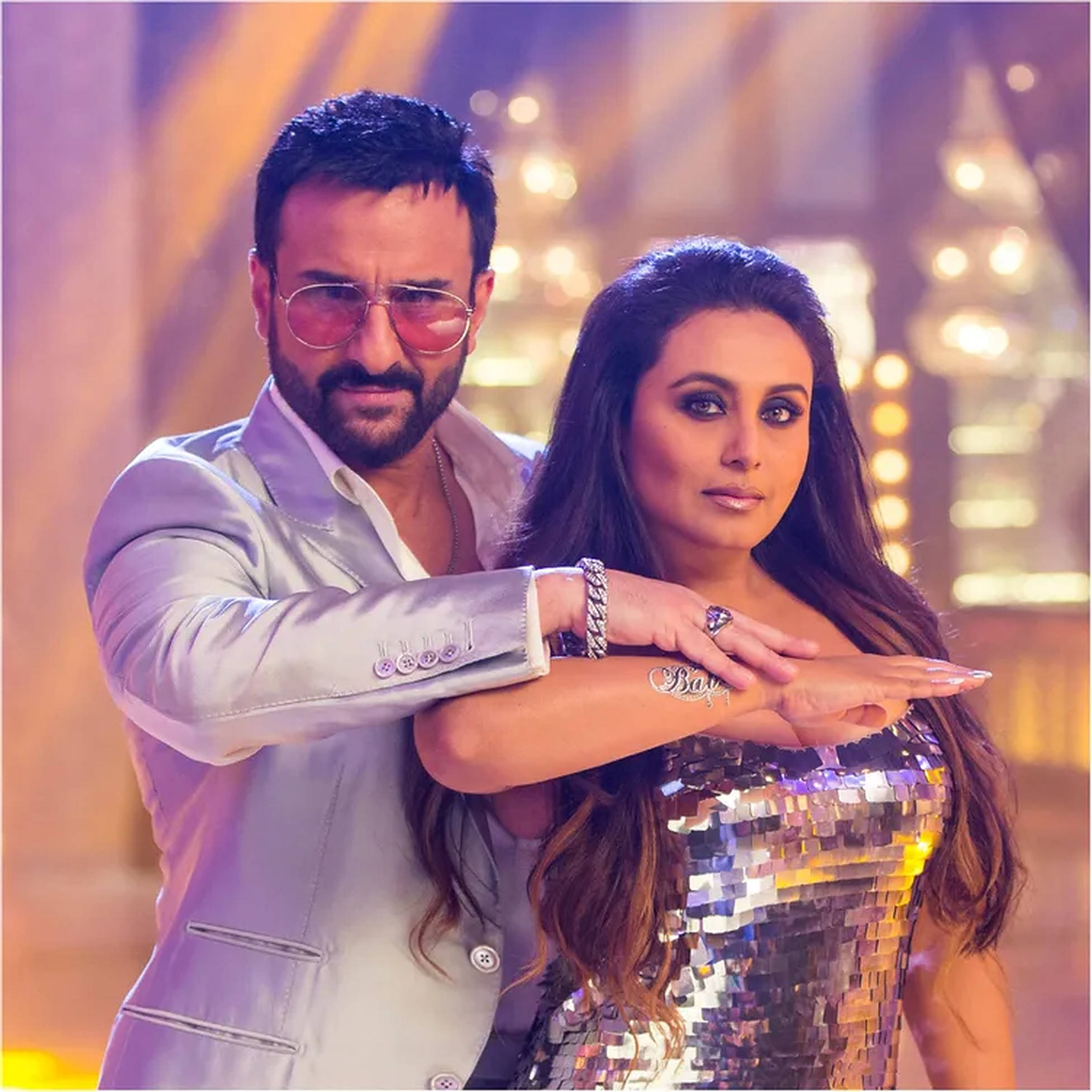 Rani Mukerji 桌布 壁紙 — 一對魅力情侶在閃爍的迪斯可場景中自信地擺姿勢，伴隨著溫暖的舞台燈光和閃亮的服裝。