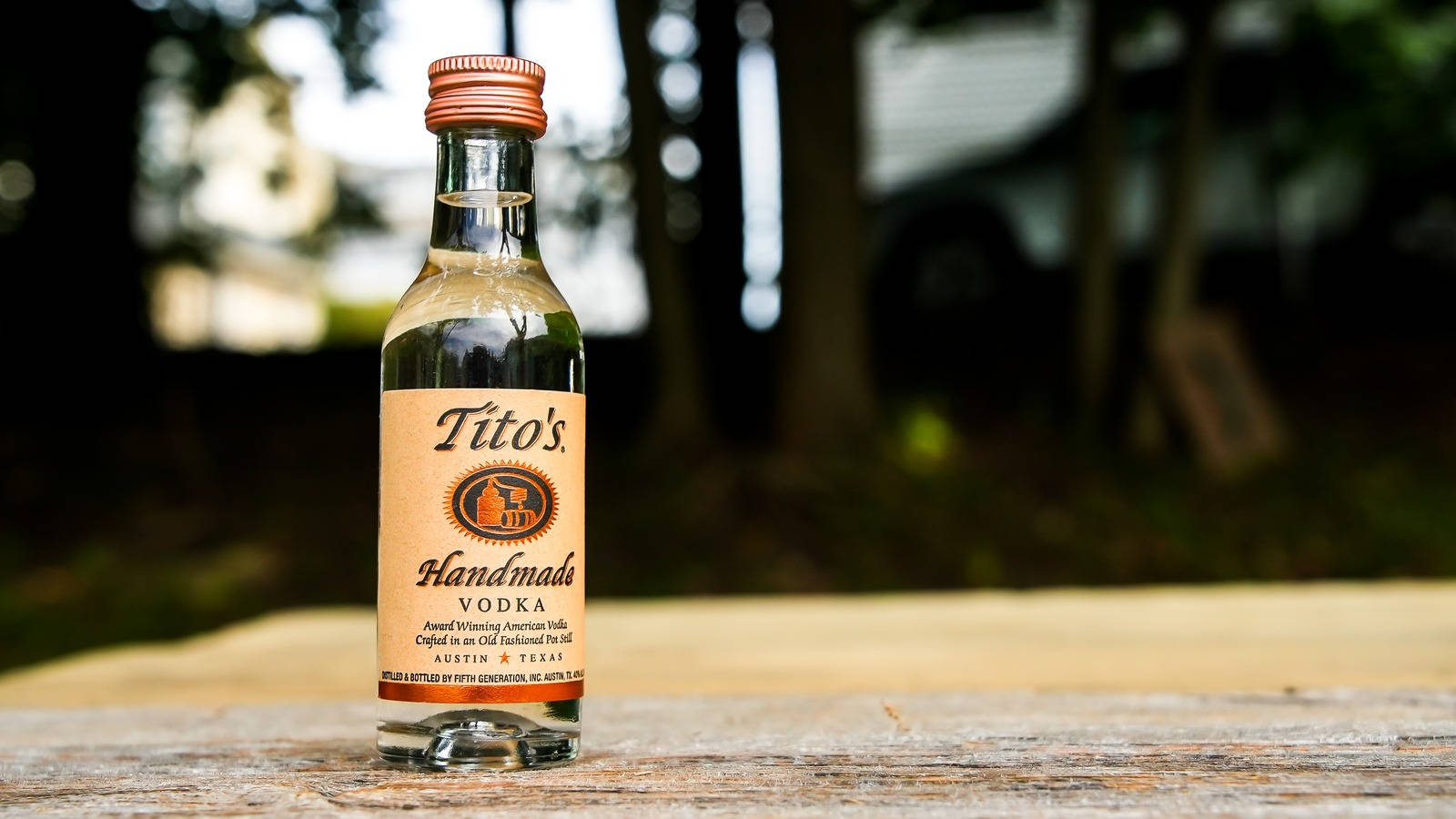 標誌 桌布 壁紙 — 一小瓶Tito's Handmade Vodka放在戶外一張風化的木桌上，背景有模糊的樹木。
