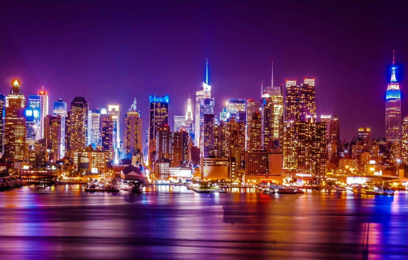 紐約市 桌布 壁紙 — 一幅令人目眩神迷的紐約夜景，摩天大樓在深紫色天空下閃耀，燈光倒映在下方漆黑的水面上。