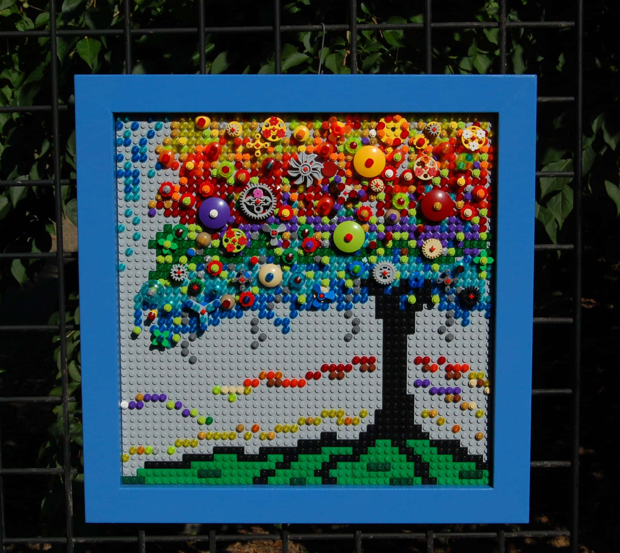 Lego Art 桌布 壁紙 — 一幅充滿活力的LEGO樹藝術品，帶有色彩繽紛的鈕扣葉片和齒輪裝飾，以藍色畫框鑲嵌在綠色籬笆背景上。