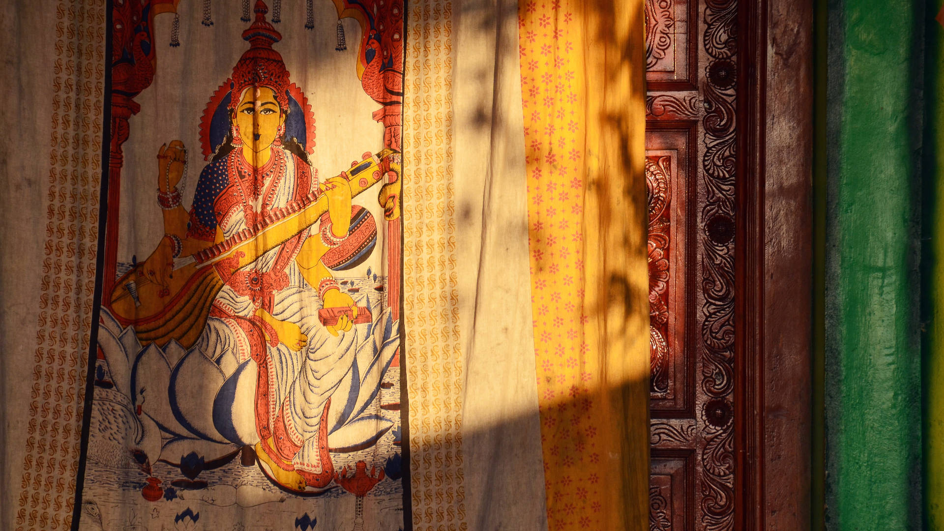 Saraswati 桌布 壁紙 — 一幅充滿活力的傳統印度神祇畫作，沐浴在透過圖案窗簾過濾的金色陽光中。