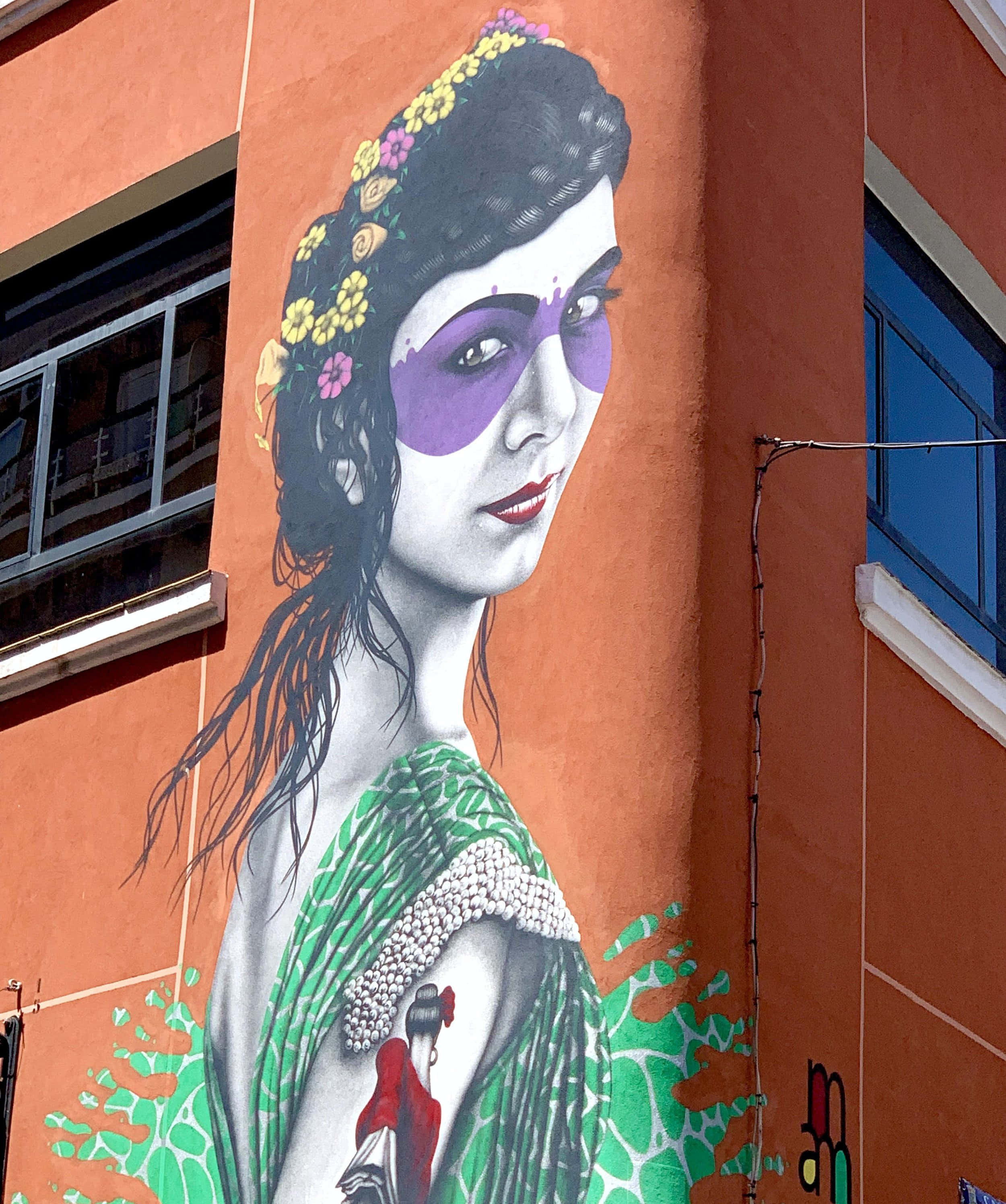 Graffiti Art 桌布 壁紙 — 一幅充滿活力的壁畫，描繪了一位戴著紫色眼罩、頭髮上點綴著花朵的女性，裝飾在橙色建築物的外牆上。