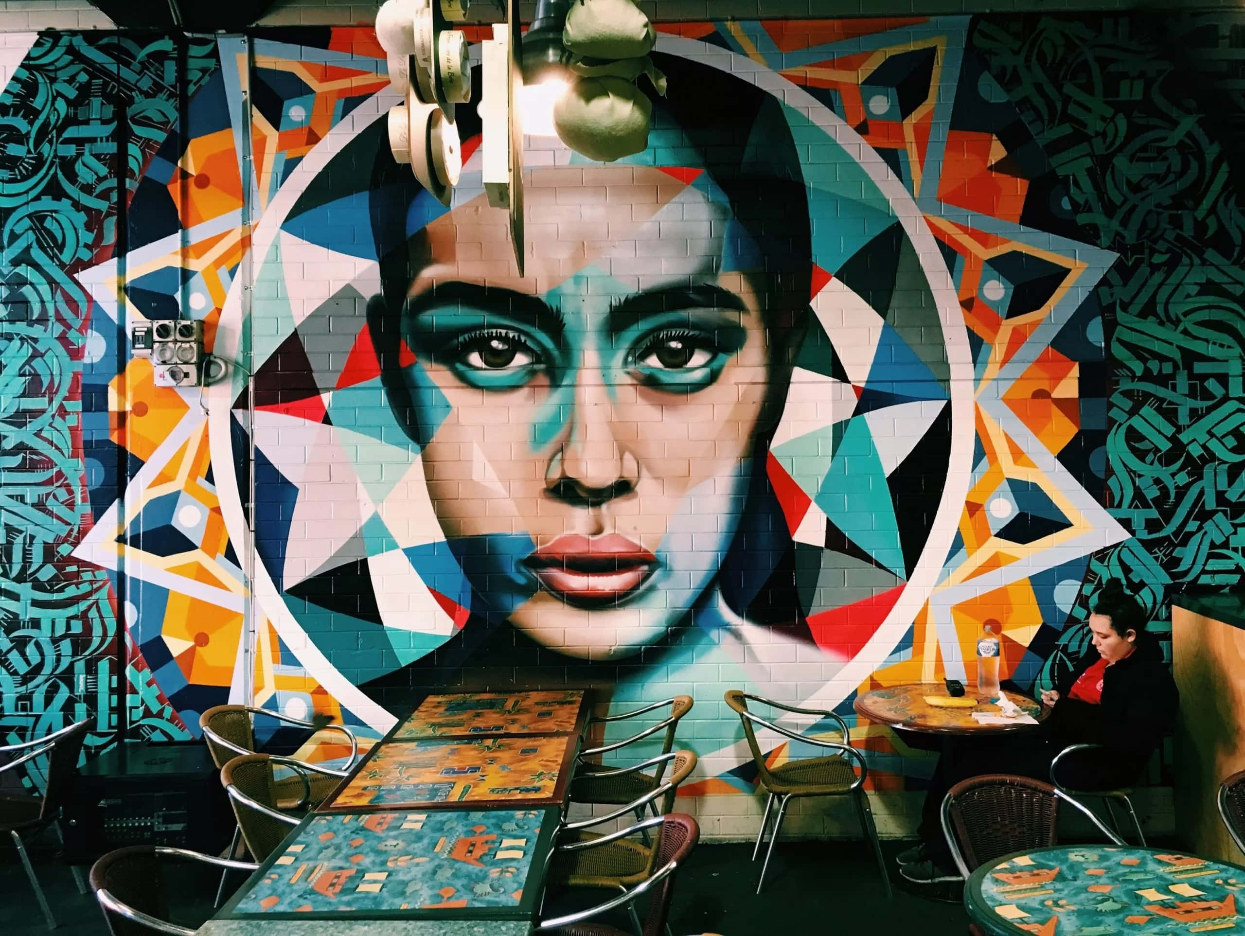 Wall Painting 桌布 壁紙 — 一幅充滿活力的幾何女性面孔壁畫主導了磚牆，在燈光昏暗的咖啡館裡，有圖案桌子和一位坐在附近的顧客。