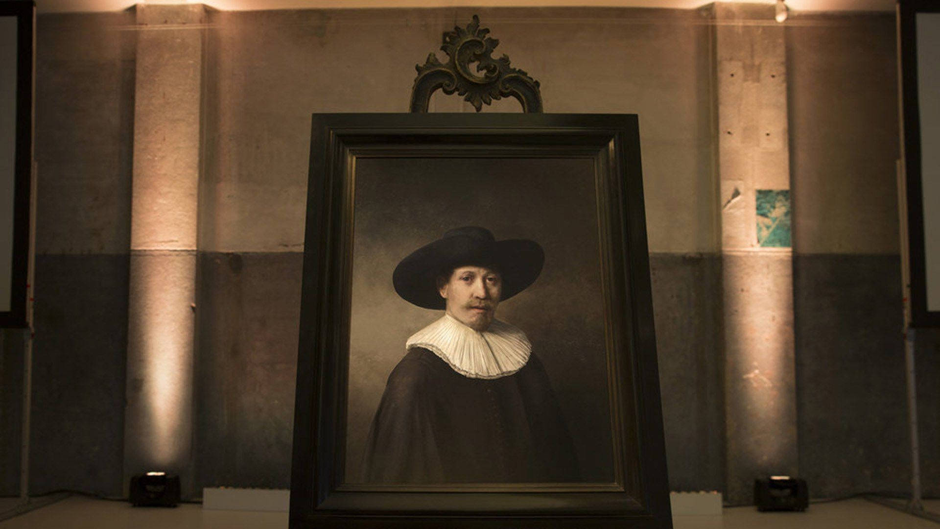 Rembrandt 桌布 壁紙 — 一幅古典肖像畫，描繪一位戴黑帽、穿白色皺領的男子，懸掛在燈光昏暗、混凝土牆壁和聚光燈的畫廊中。