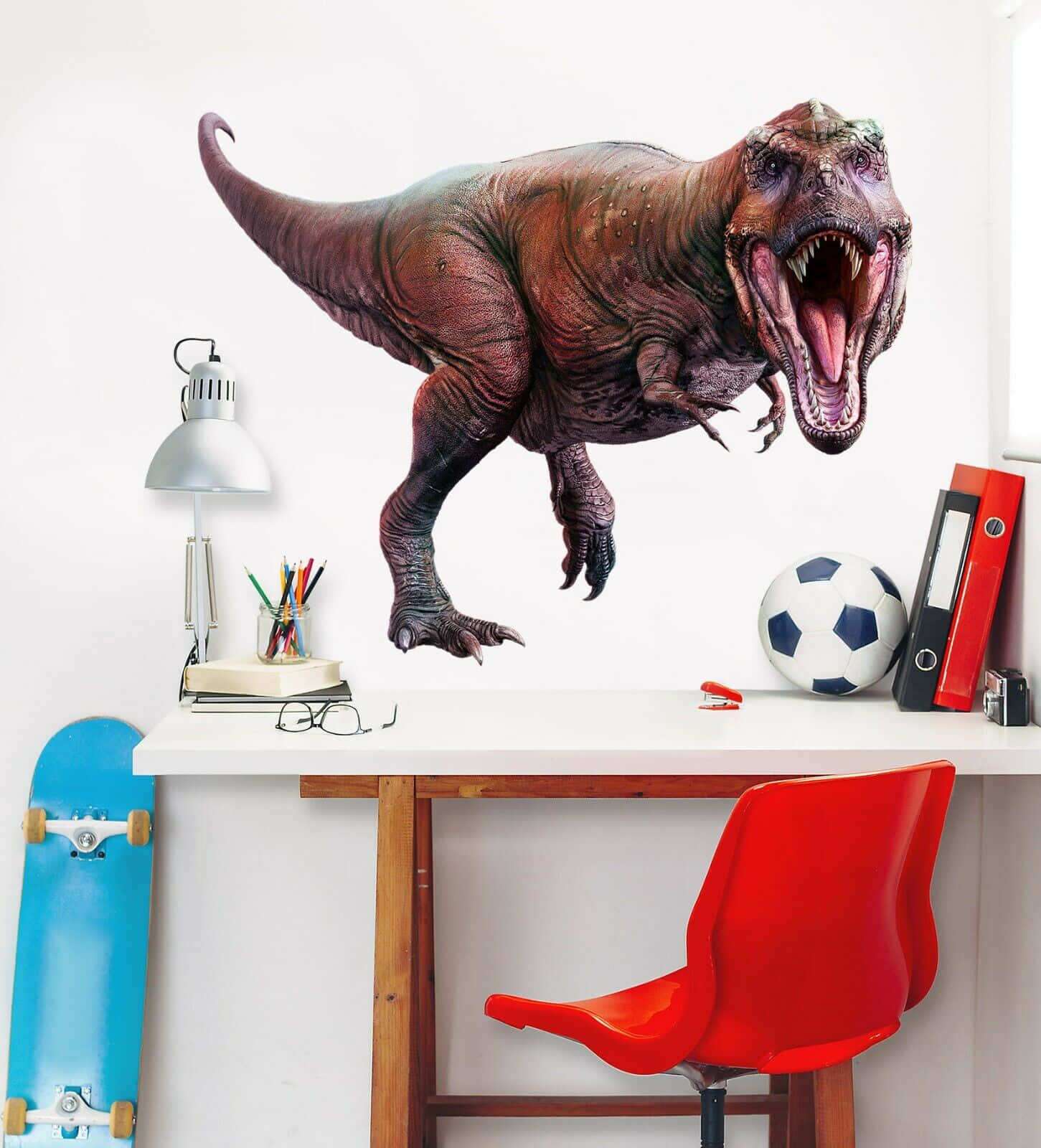 3d Dinosaur 桌布 壁紙 — 一幅咆哮的暴龍壁紙主宰了兒童書房的牆壁，牆下是一張書桌，上面有紅色椅子、足球和滑板。