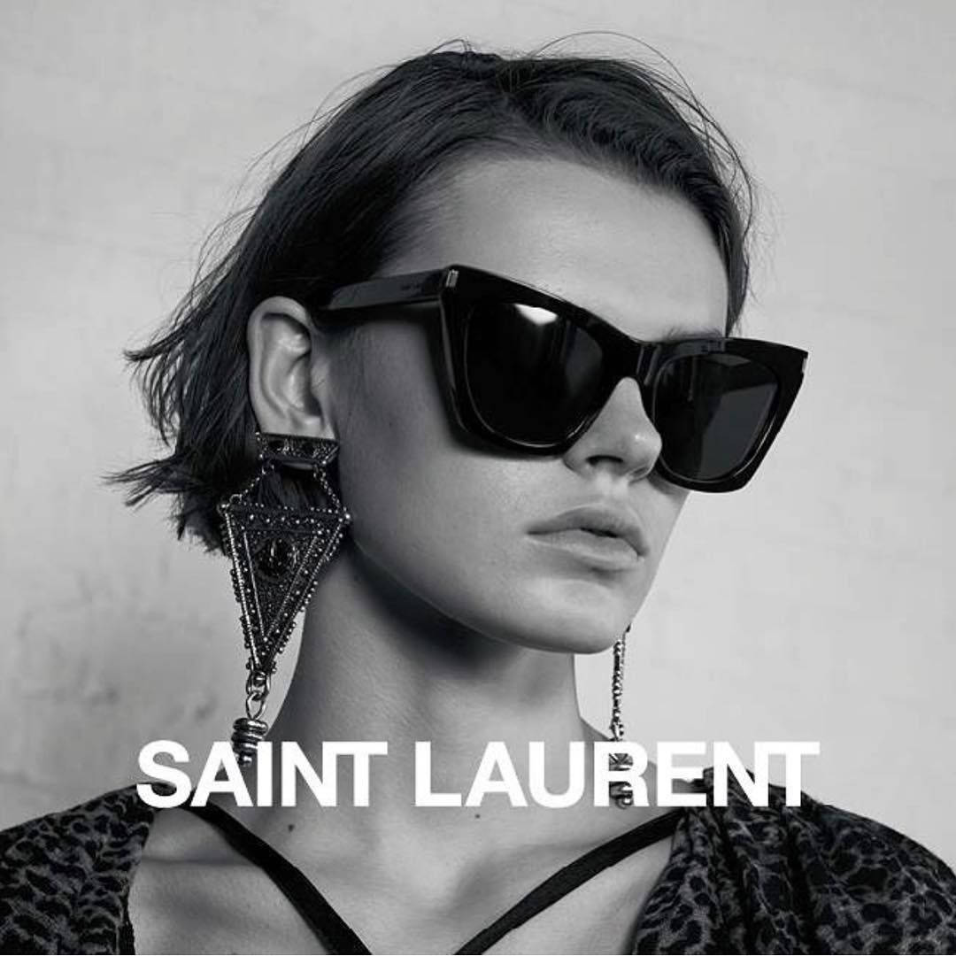 Ysl 桌布 壁紙 — 一幅單色肖像，短黑髮女性戴著大膽太陽眼鏡和華麗耳環，背景為有紋理的牆壁。