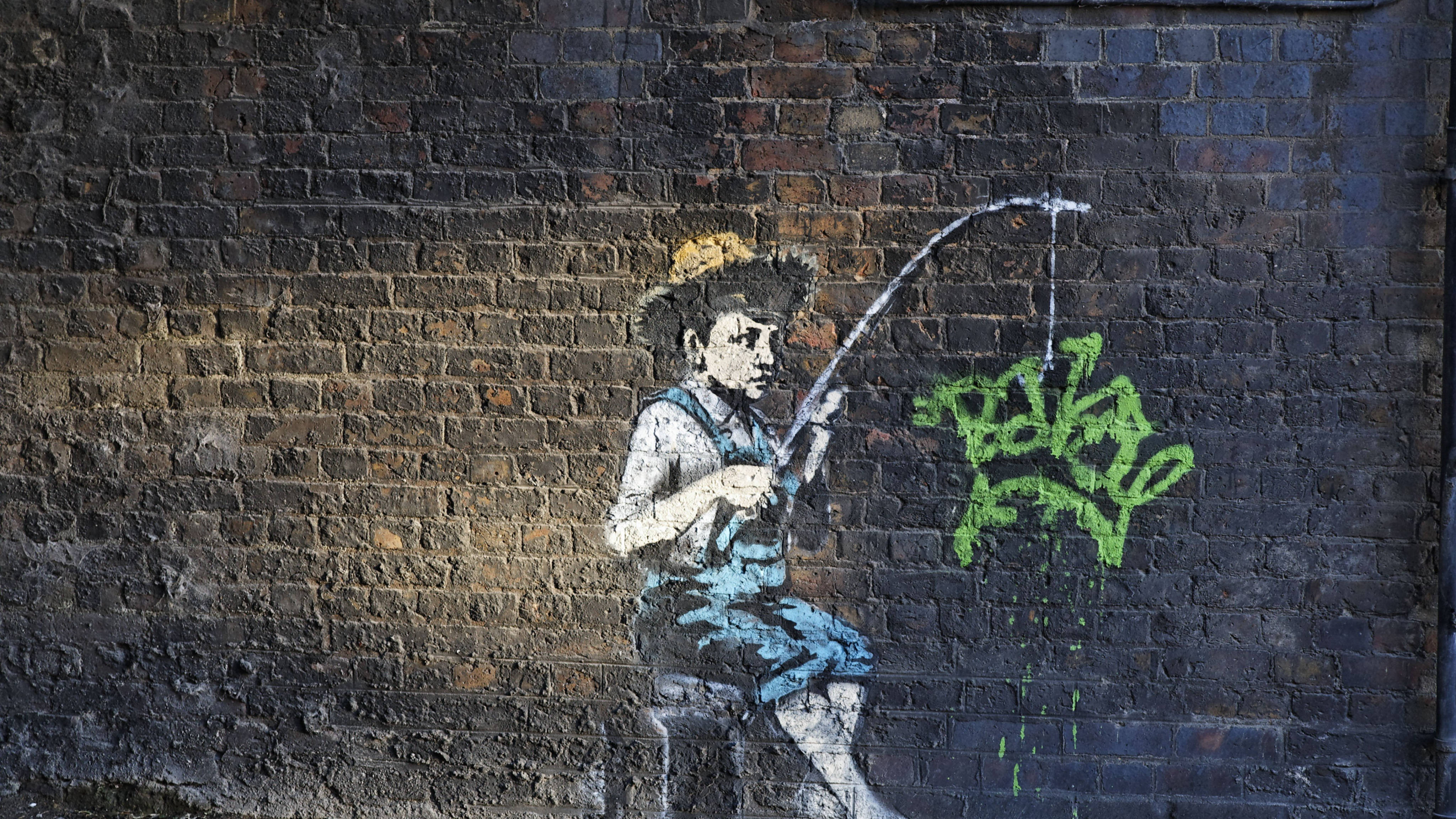 Banksy 桌布 壁紙 — 一幅塗鴉壁畫，描繪一個男孩用彎曲的釣竿釣魚，背景是風化的磚牆，附近有綠色噴漆標籤。