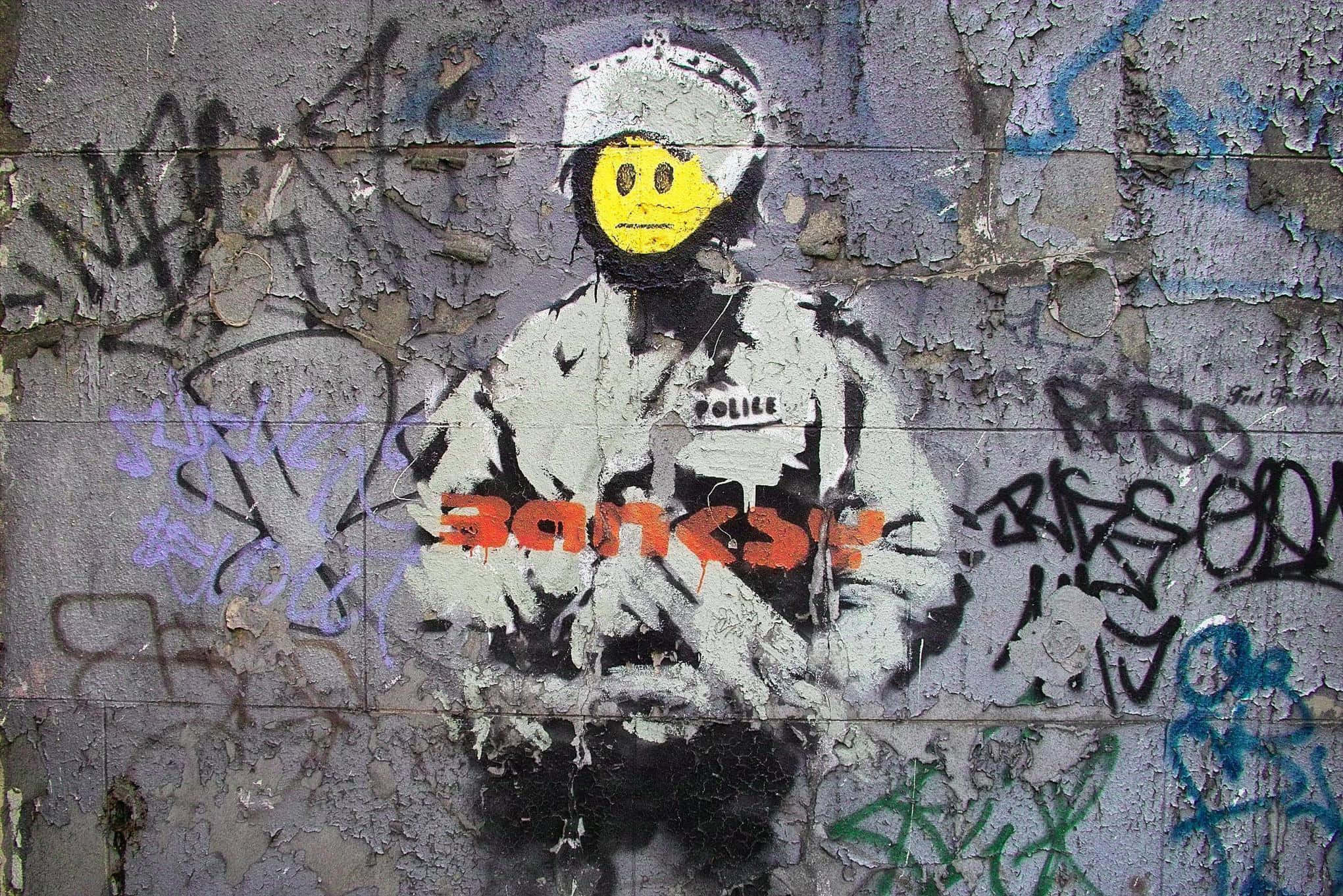 Banksy 桌布 壁紙 — 一幅塗鴉壁畫，描繪一名戴著黃色笑臉頭盔的警察，背景是佈滿塗鴉標籤的風化牆壁。