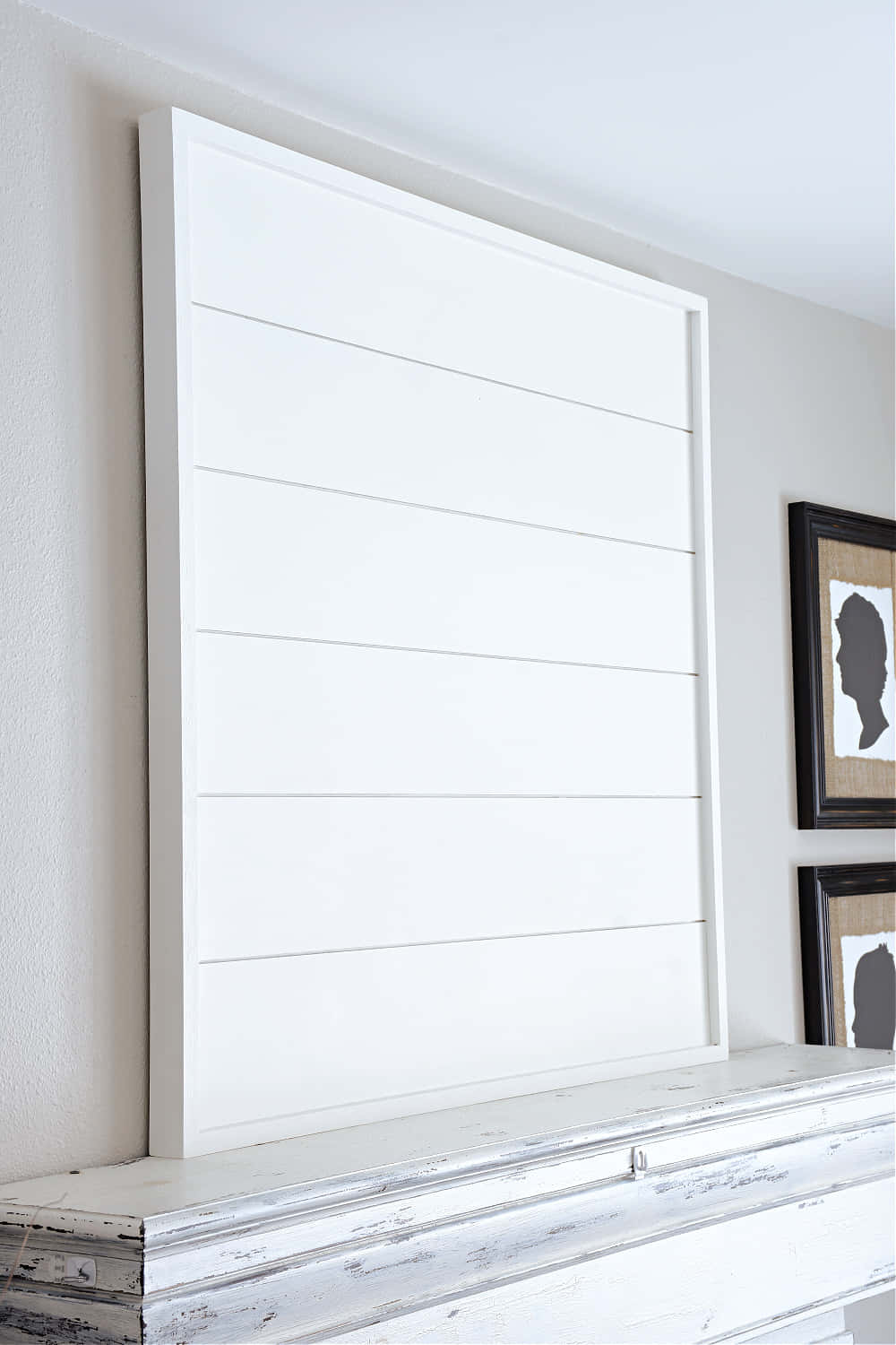 Shiplap 桌布 壁紙 — 一幅大型白色船舷板風格畫框的藝術品靠在淺灰色牆上，下方是一個殘舊的白色壁爐架，旁邊有兩個黑色畫框的剪影肖像。