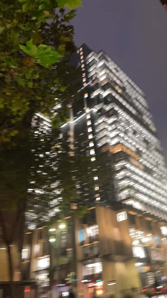 Building 桌布 壁紙 — 一幅帶動態模糊效果的夜間城市景觀，展示一座高聳的玻璃摩天大樓，亮著燈的窗戶被茂密的樹枝框住。