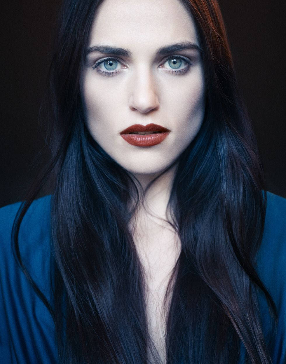 Katie 桌布 壁紙 — 一幅引人注目的女性肖像，擁有強烈的藍色眼睛和長長的深色頭髮，背景為深色。
