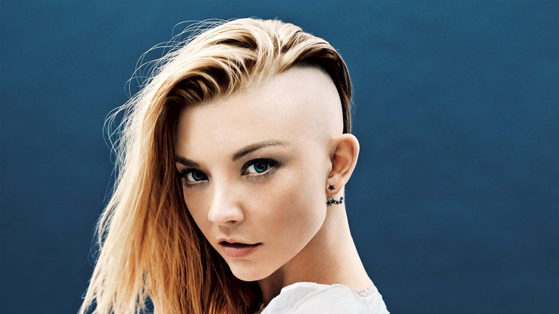 natalie dormer 桌布 壁紙 — 一幅引人注目的肖像，一位年輕女性擁有大胆的金色漸層髮型和銳利的藍色眼睛，背景為深藍色。