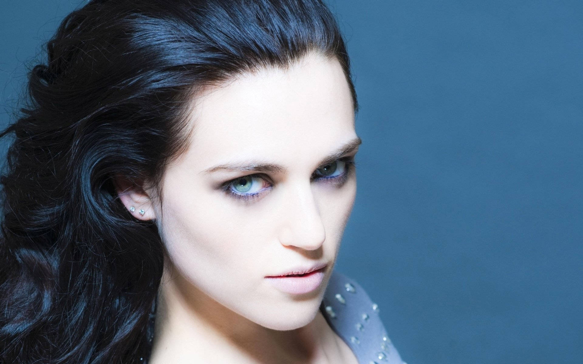 Katie Mcgrath 桌布 壁紙 — 一幅引人注目的肖像，一位擁有強烈藍色眼睛和深色頭髮的女性，背景是冷藍色。
