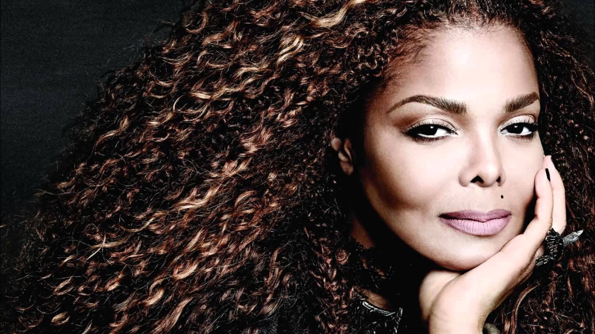 janet jackson 桌布 壁紙 — 一幅引人注目的肖像，一位擁有豐盈棕色捲髮的女性，在深色背景下將下巴擱在手上。