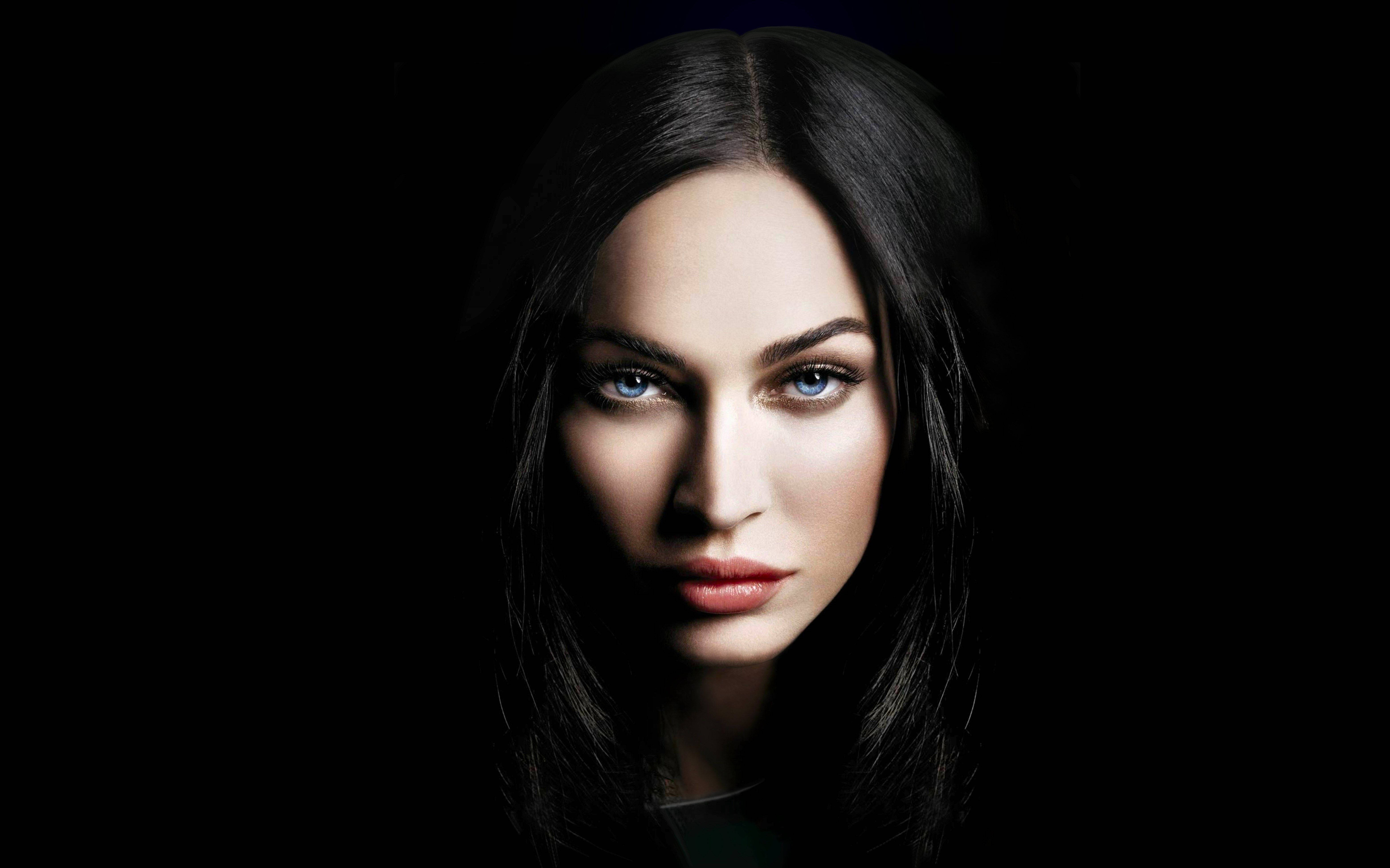 Megan Fox 桌布 壁紙 — 一幅引人注目的肖像，一位擁有銳利藍眼睛和深色頭髮的女性，背景為純黑色。