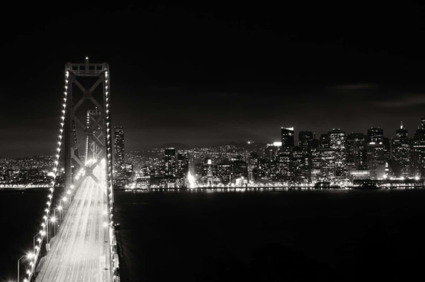 Cool Black And White 桌布 壁紙 — 一幅引人注目的黑白夜景，展示一座燈火通明的吊橋，通往水面上閃爍的城市天際線。