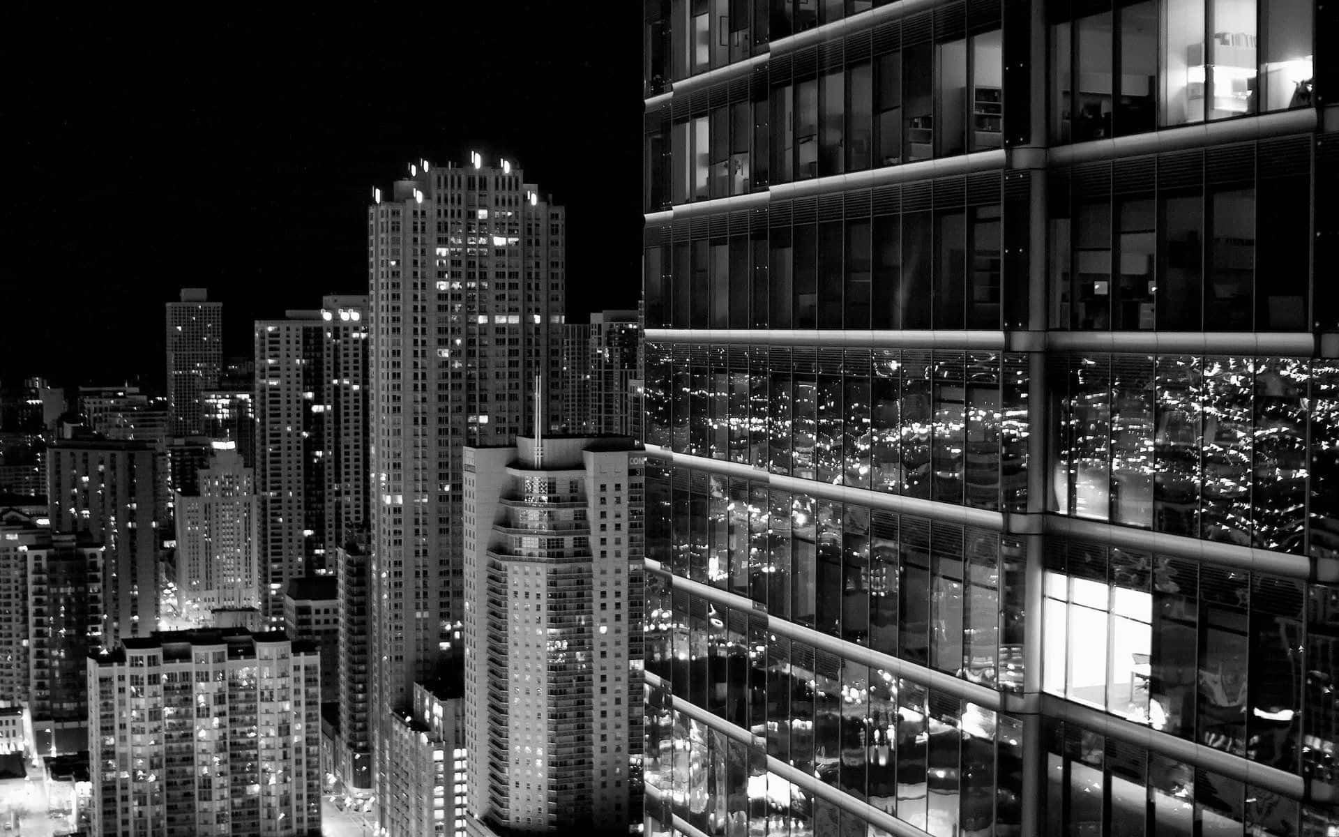Black And White Cityscape 桌布 壁紙 — 一幅引人注目的黑白夜景城市景觀，展示高聳的摩天大樓，其明亮的窗戶反射出充滿活力的城市燈光。