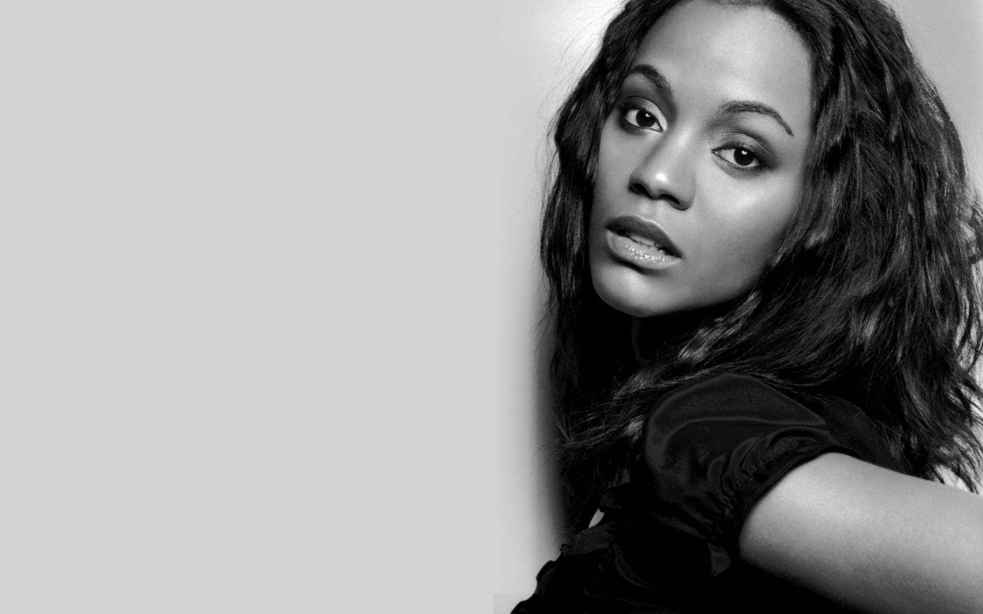 Zoe Saldana 桌布 壁紙 — 一幅引人注目的黑白肖像，一位長黑髮的女性在淺灰色純色背景前直視鏡頭。
