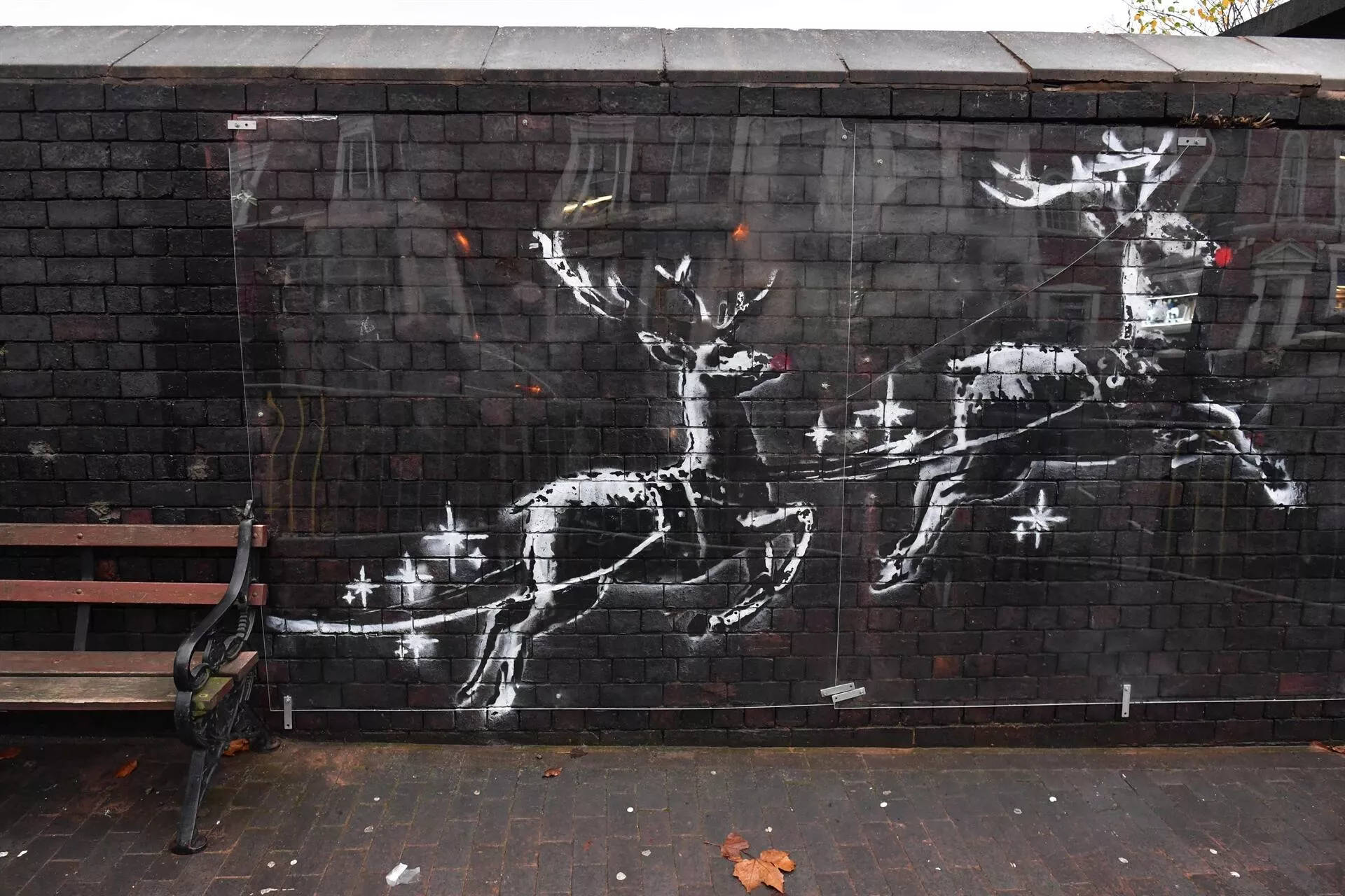 Banksy 桌布 壁紙 — 一幅引人注目的黑白街頭壁畫，描繪馴鹿拉著雪橇，雪橇上有星星，位於深色磚牆旁，旁邊有一張木長椅。