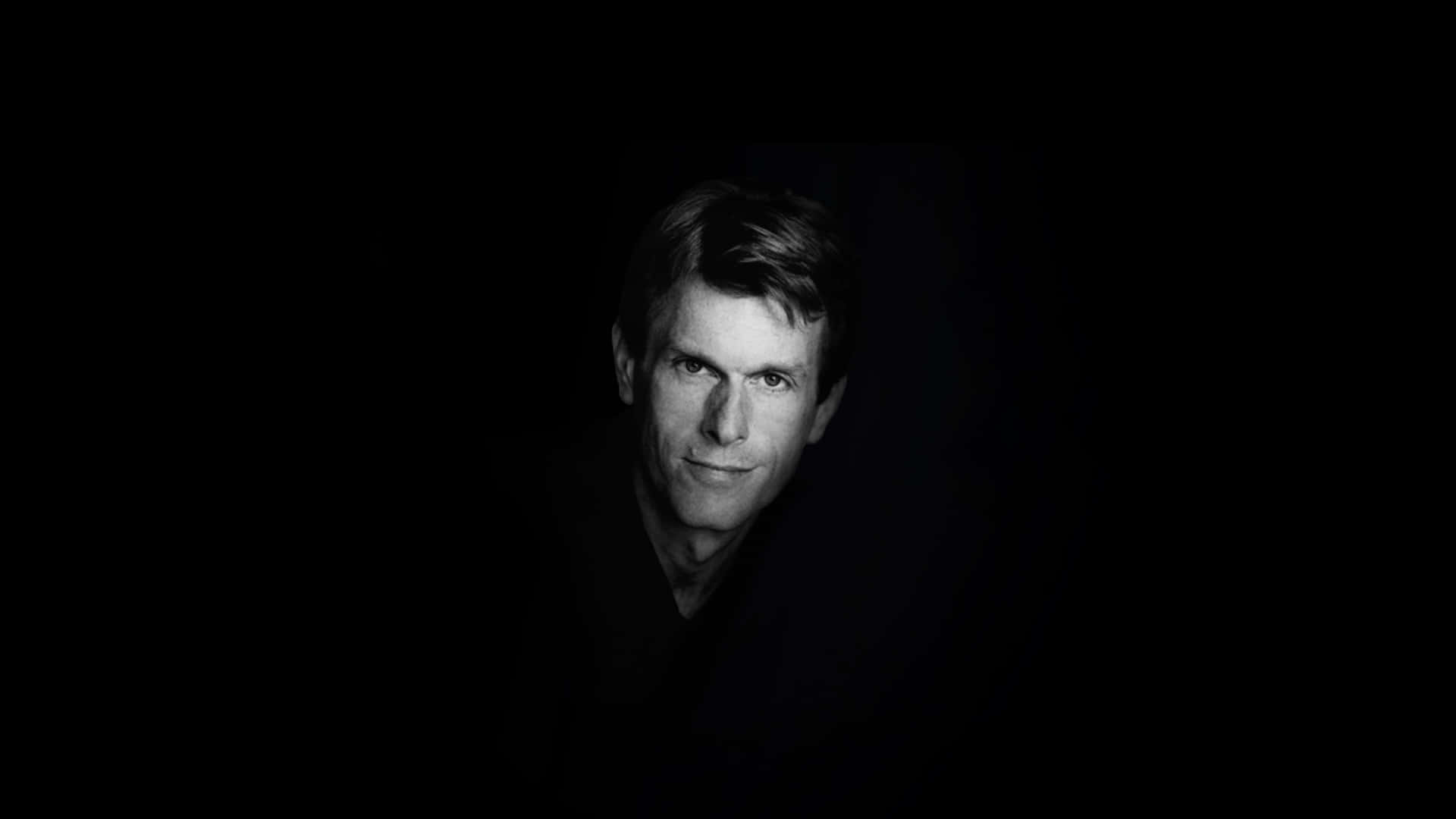 Kevin Conroy 桌布 壁紙 — 一幅強烈黑白肖像，捕捉一位眼神銳利、帶著微妙微笑的男子，背景完全漆黑。