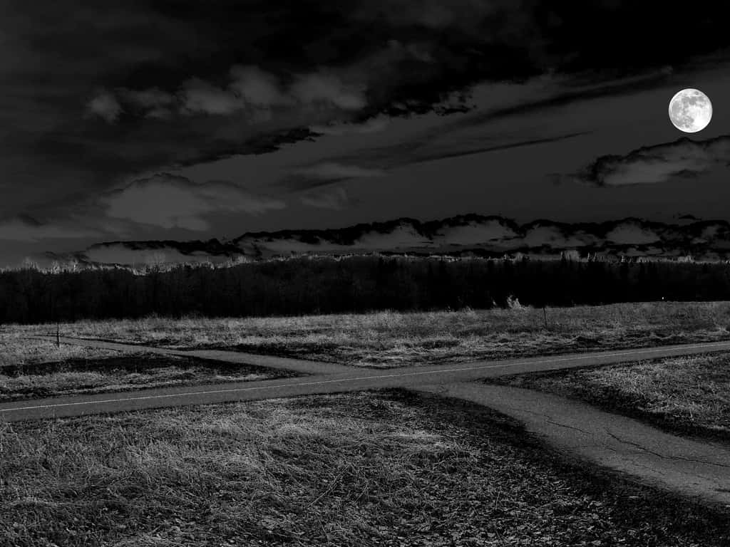 Dark Crossroad 桌布 壁紙 — 一幅清晰的黑白夜景，顯示一條月光照耀的道路穿過田野，遠處有山丘，天空多雲。