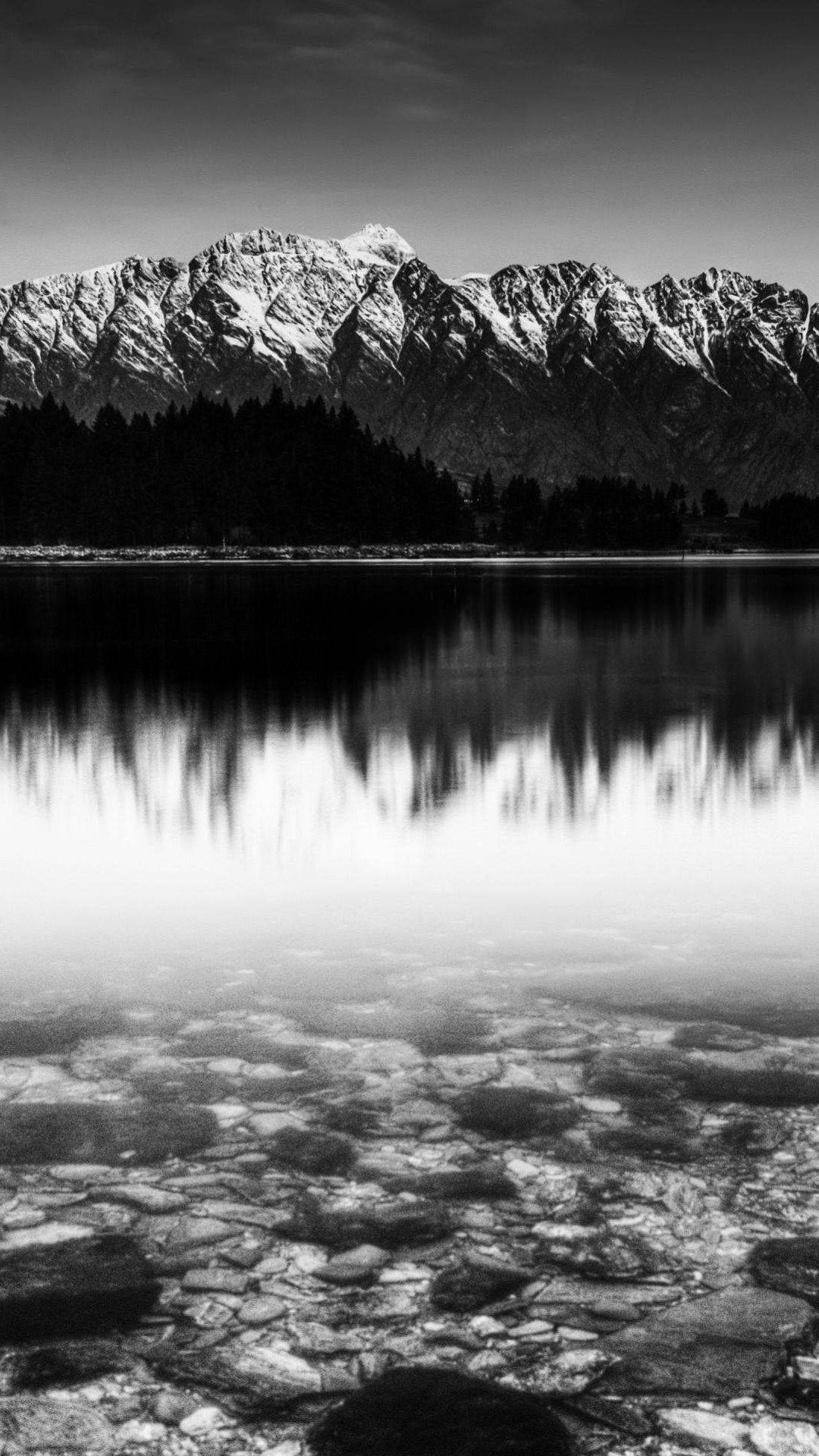 積雪覆蓋的山脈倒映在平靜的湖面上，淺水中可見鵝卵石。 — 黑白美學動圖 壁紙