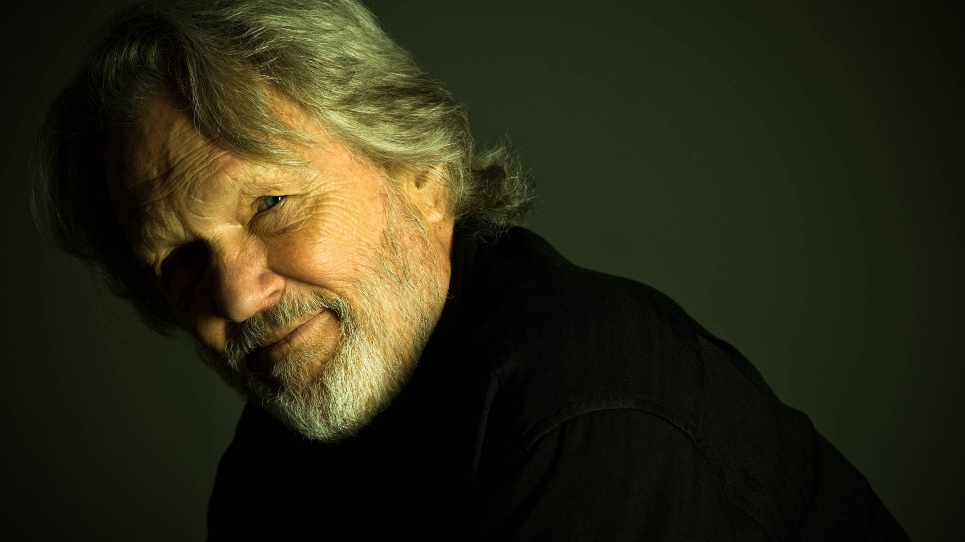 Kris Kristofferson 桌布 壁紙 — 一幅溫暖燈光的肖像，一位灰白頭髮和鬍鬚的老年男性在深色背景前溫柔微笑。