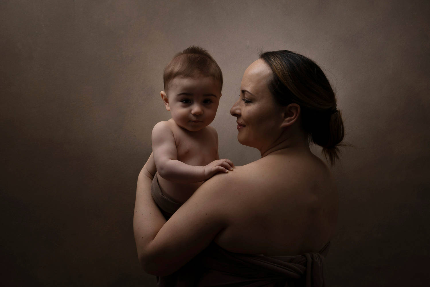 Mother And Baby 桌布 壁紙 — 一幅溫柔的肖像，母親在柔和的棕色背景和溫暖燈光下抱著她的嬰兒。