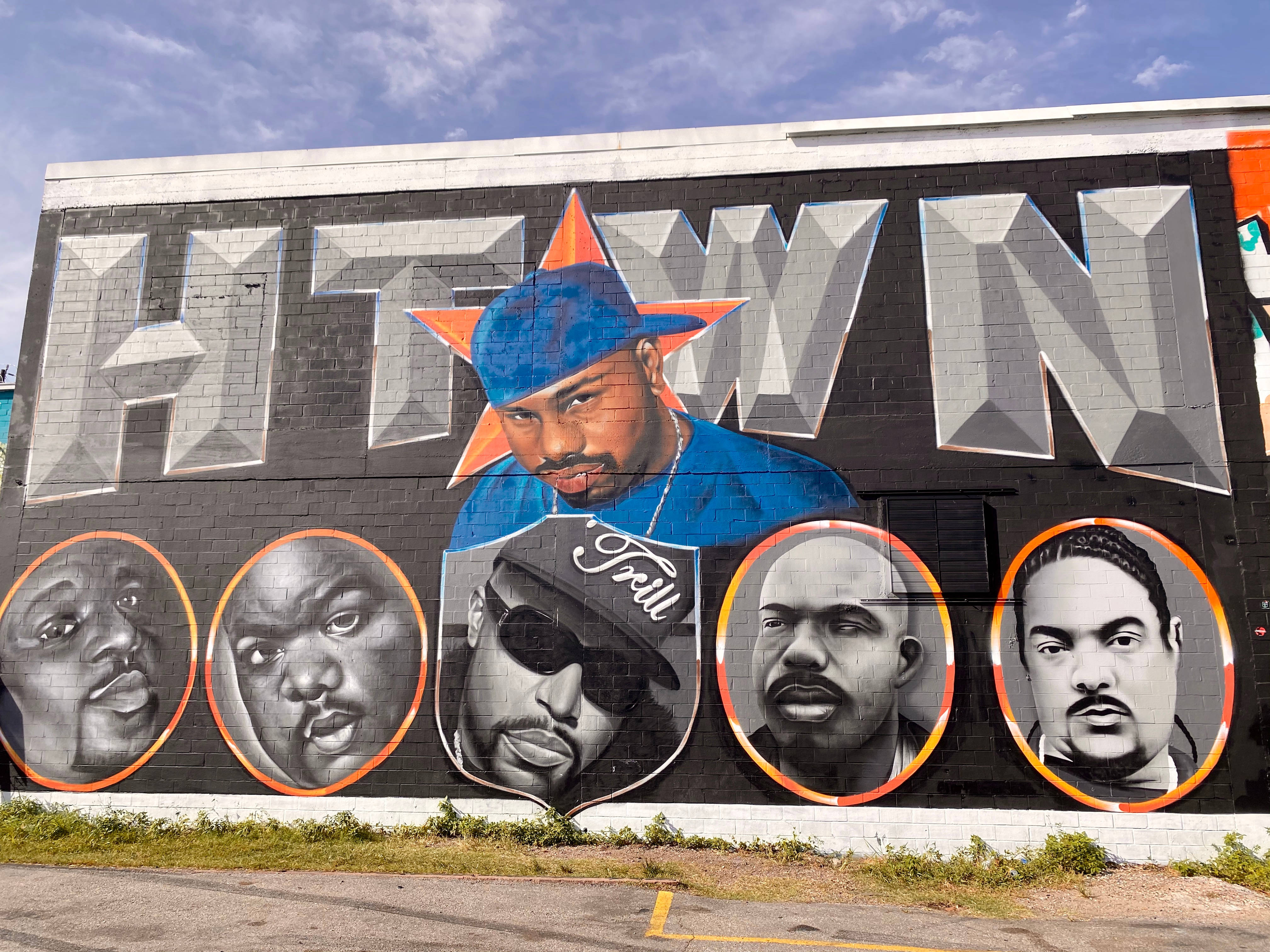 Houston 桌布 壁紙 — 一幅磚牆上的鮮豔塗鴉壁畫，上面有「HTWN」字樣，中央是一位戴藍色帽子和星星的男士肖像，兩側是四位標誌性說唱歌手的圓形肖像，背景是晴朗的藍天。