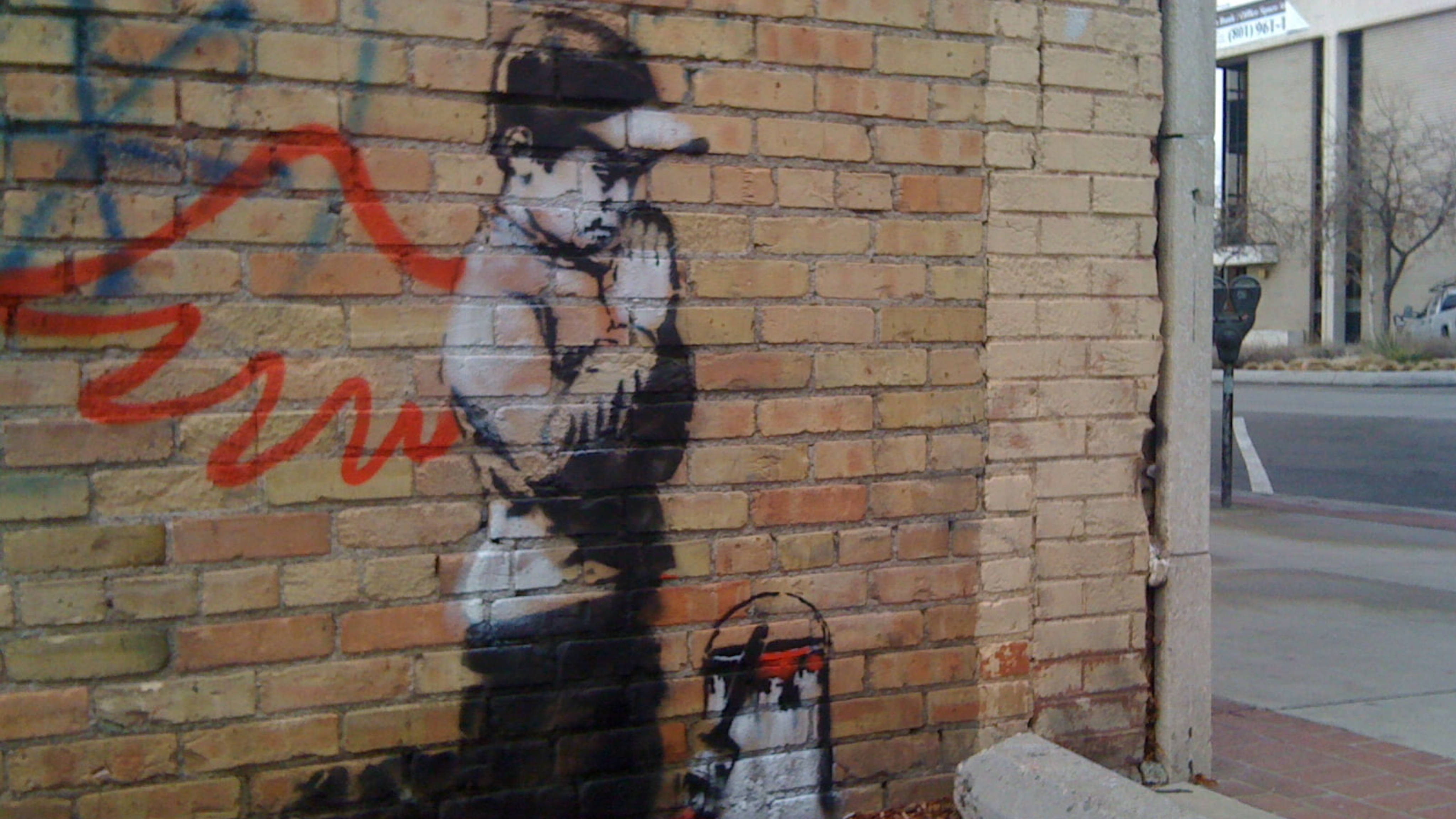 Banksy 桌布 壁紙 — 一幅粗糙的街頭壁畫，描繪一個戴帽子和手套的男孩，畫在人行道旁風化的磚牆上。