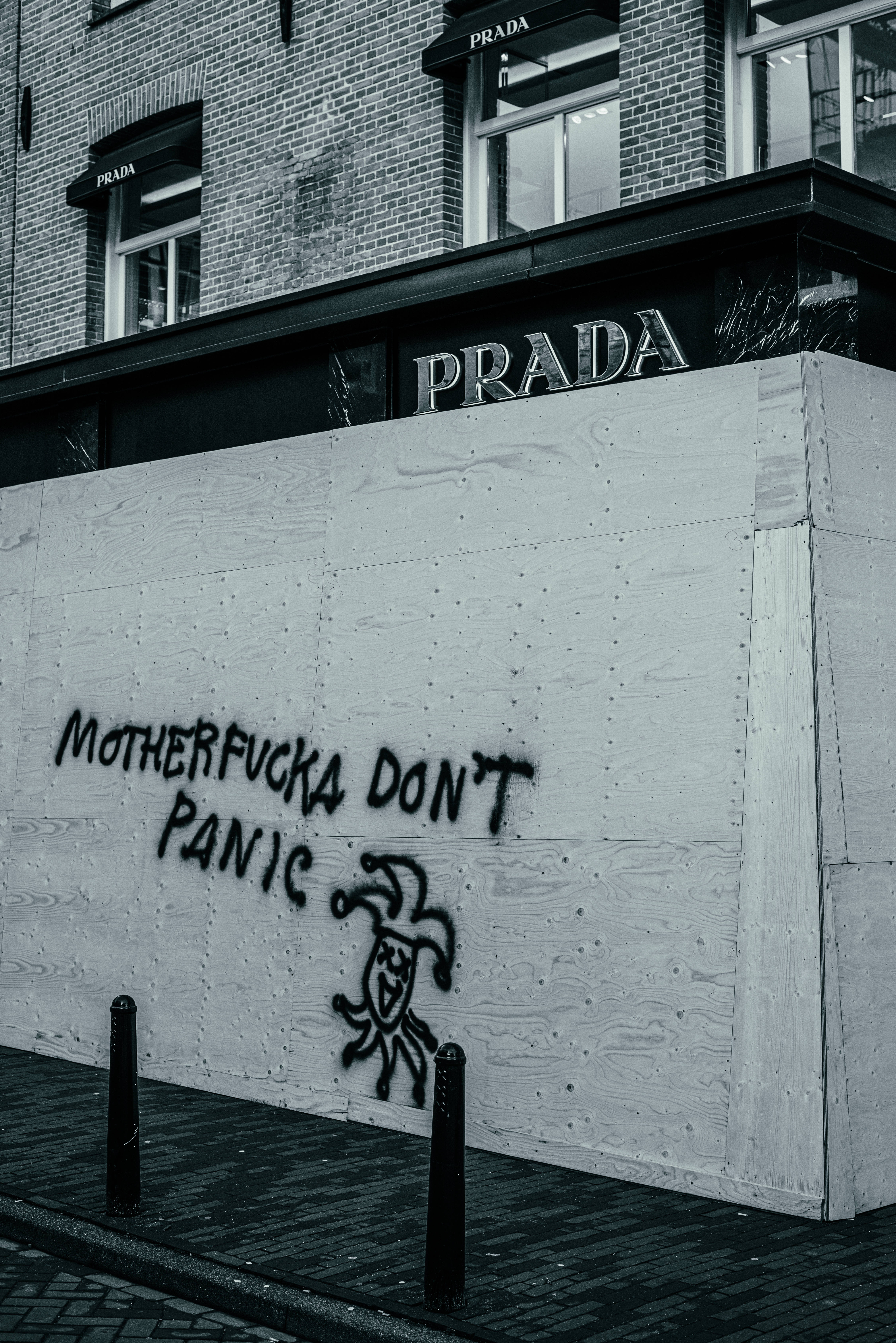 Prada 桌布 壁紙 — 一幅粗糙的黑白城市場景，顯示Prada商店的外牆部分被膠合板牆遮擋，牆上塗有粗體塗鴉，寫著「MOTHERFUCKA DON'T PANIC」和一個小丑圖案。
