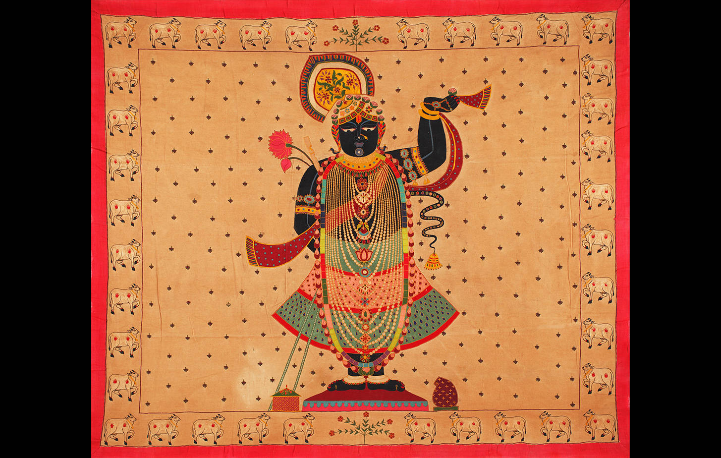 Shrinathji 桌布 壁紙 — 一幅細節豐富的傳統印度紡織藝術品，描繪了一位深色皮膚的神祇，佩戴珠寶，手持海螺和蓮花，背景為帶圖案的米色，並以紅色和牛圖案框住。