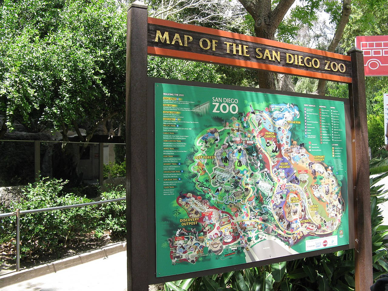 Map 桌布 壁紙 — 一幅色彩繽紛的聖地牙哥動物園插圖地圖，在明亮的陽光下，矗立在戶外，周圍環繞著茂密的綠色樹木和樹葉。