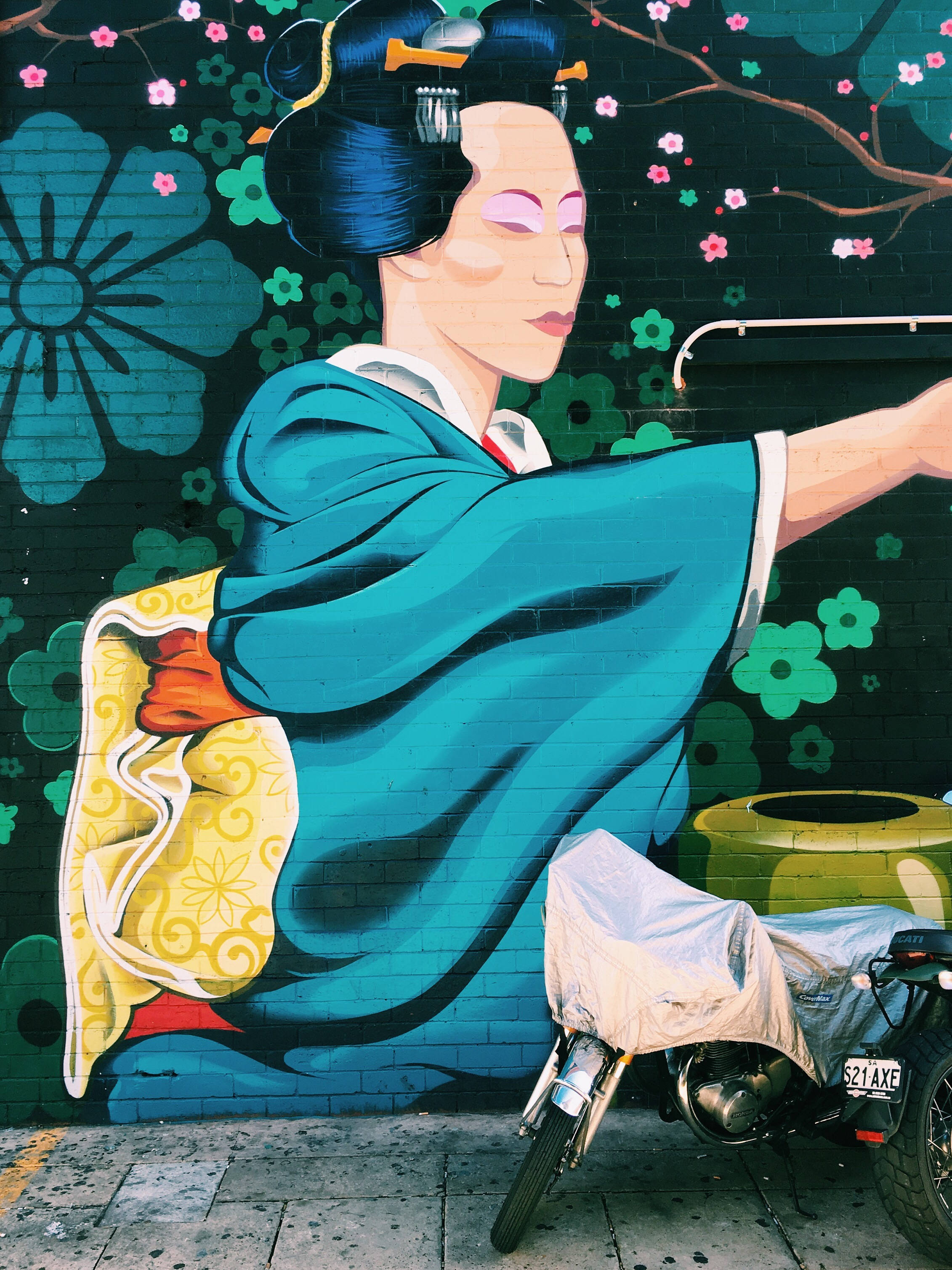 非洲藝術 桌布 壁紙 — 一幅色彩鮮豔的壁畫，描繪一位身穿藍色和服的藝妓，背景是花卉磚牆，下方停著一輛機車。