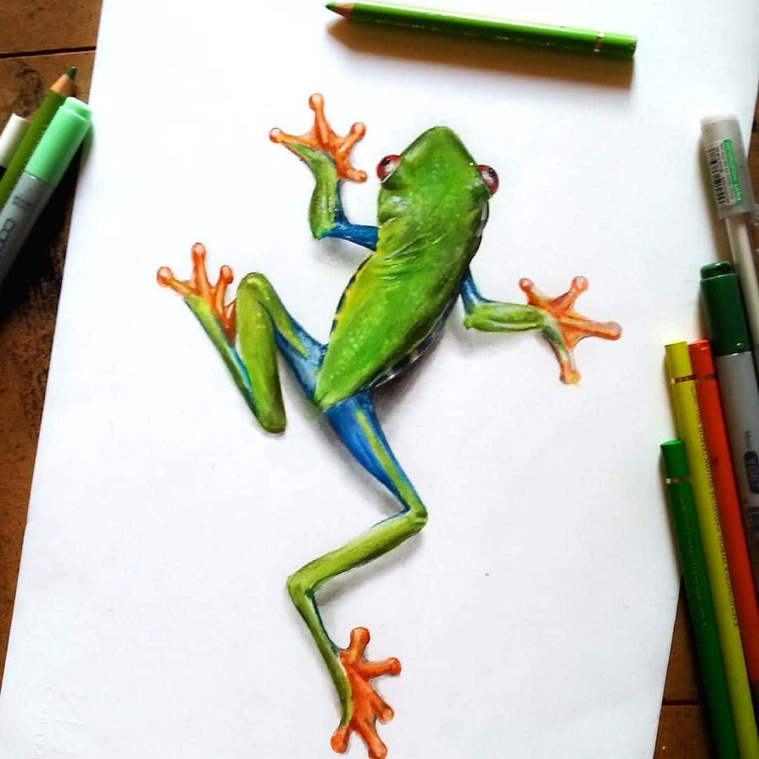 Frog Drawing 桌布 壁紙 — 一幅色彩鮮豔的青蛙畫作，橙色腳趾和藍色腿，看似要從被美術用品環繞的白紙上跳出來。