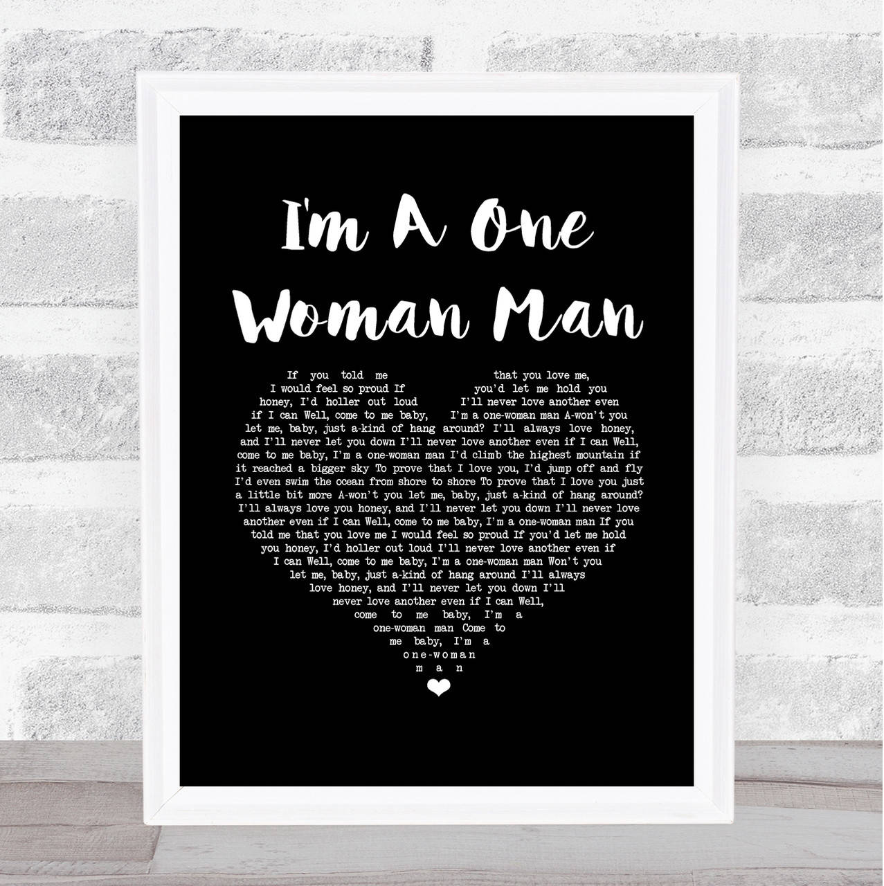 鄉村 桌布 壁紙 — 一幅裱框的黑白歌詞海報，為「I'm A One Woman Man」而設，靠在一面有紋理的灰色磚牆上，置於木地板上。