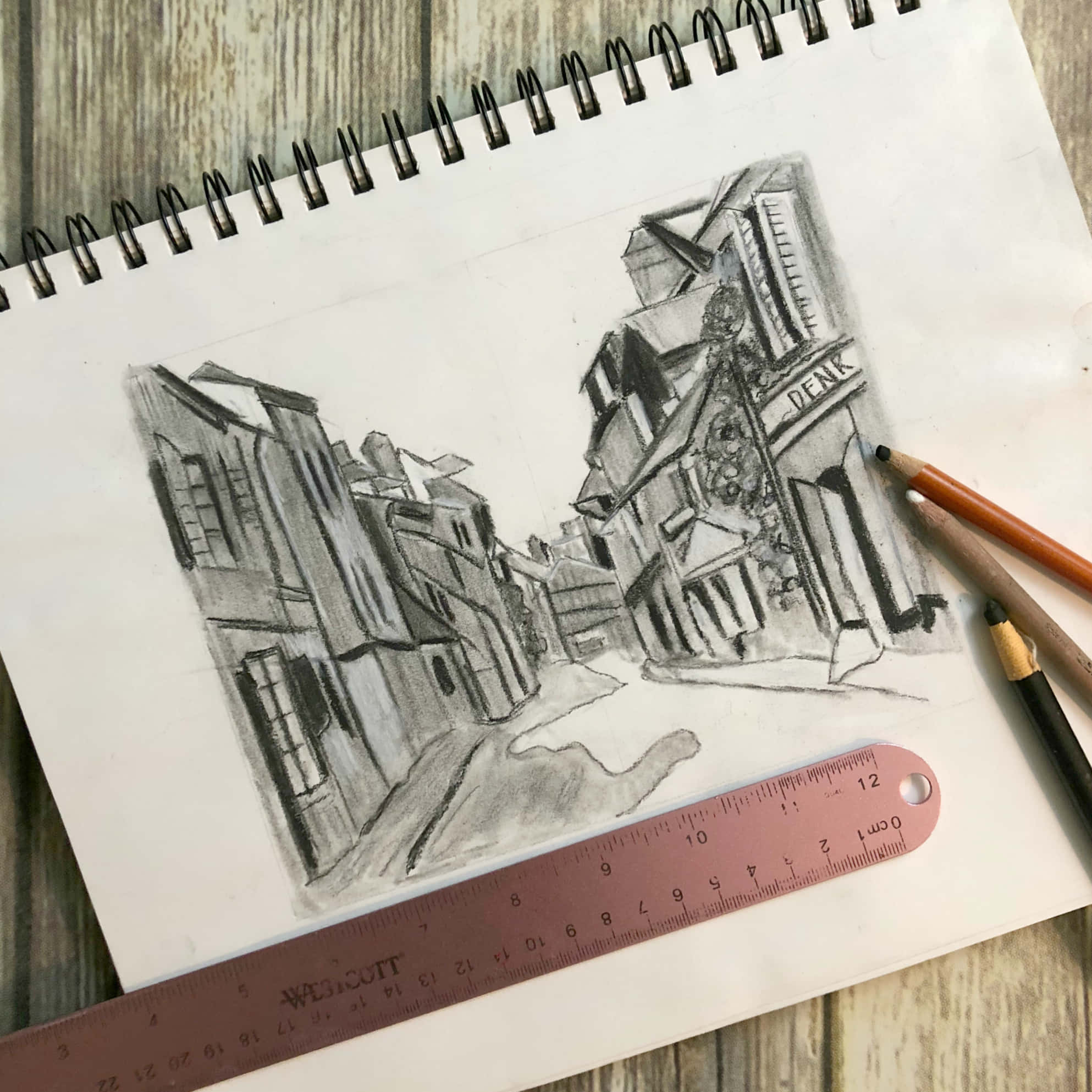 Sketch Art 桌布 壁紙 — 一幅詳細的鉛筆素描，描繪了一條狹窄的街道，有建築物和一個「DEAK」標誌，放在木質表面上，旁邊有鉛筆和一把尺子。
