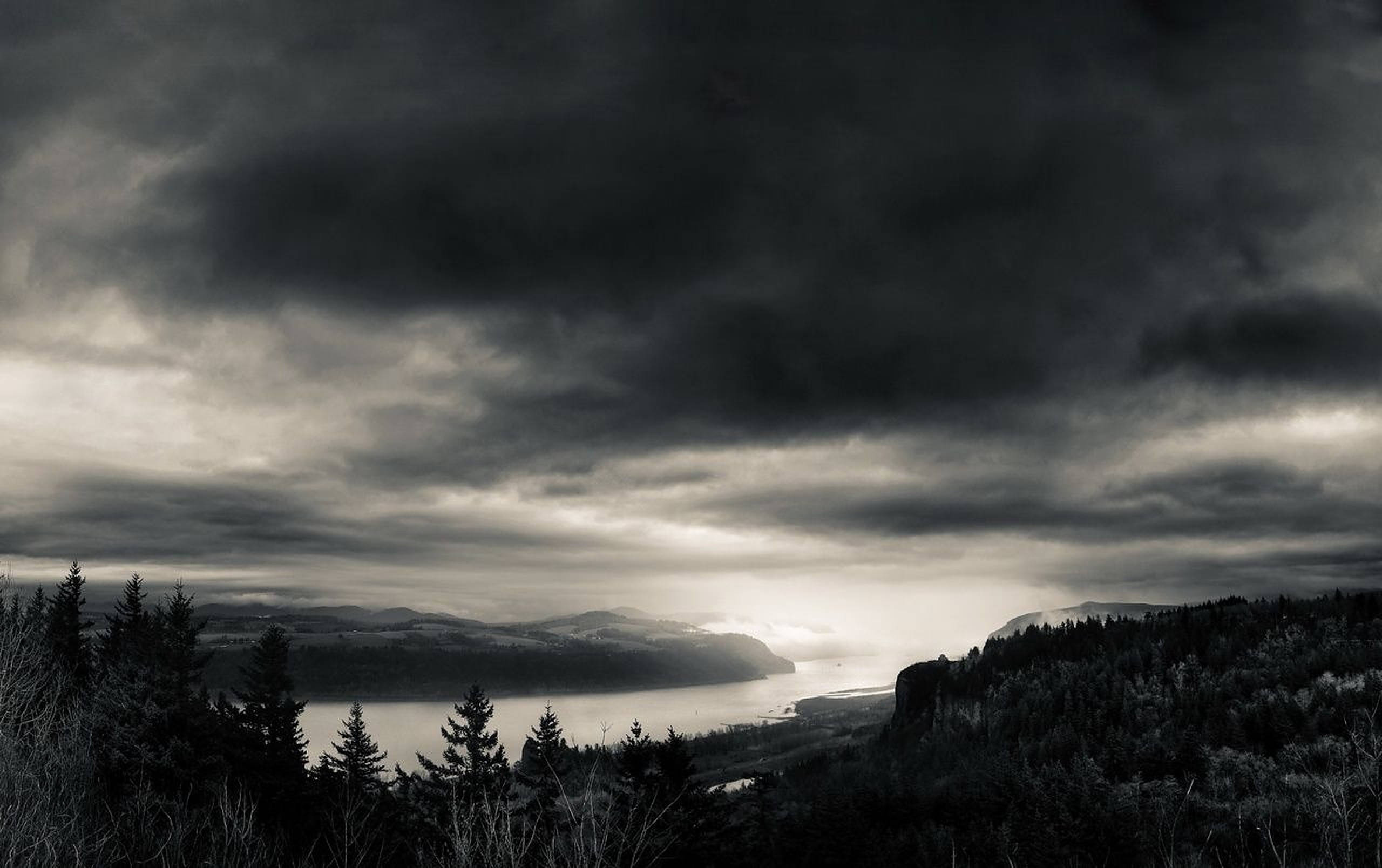 Weather 桌布 壁紙 — 一幅陰鬱的黑白風景畫，展示了一個霧氣瀰漫的湖泊，坐落在森林覆蓋的山丘之間，天空烏雲密布。