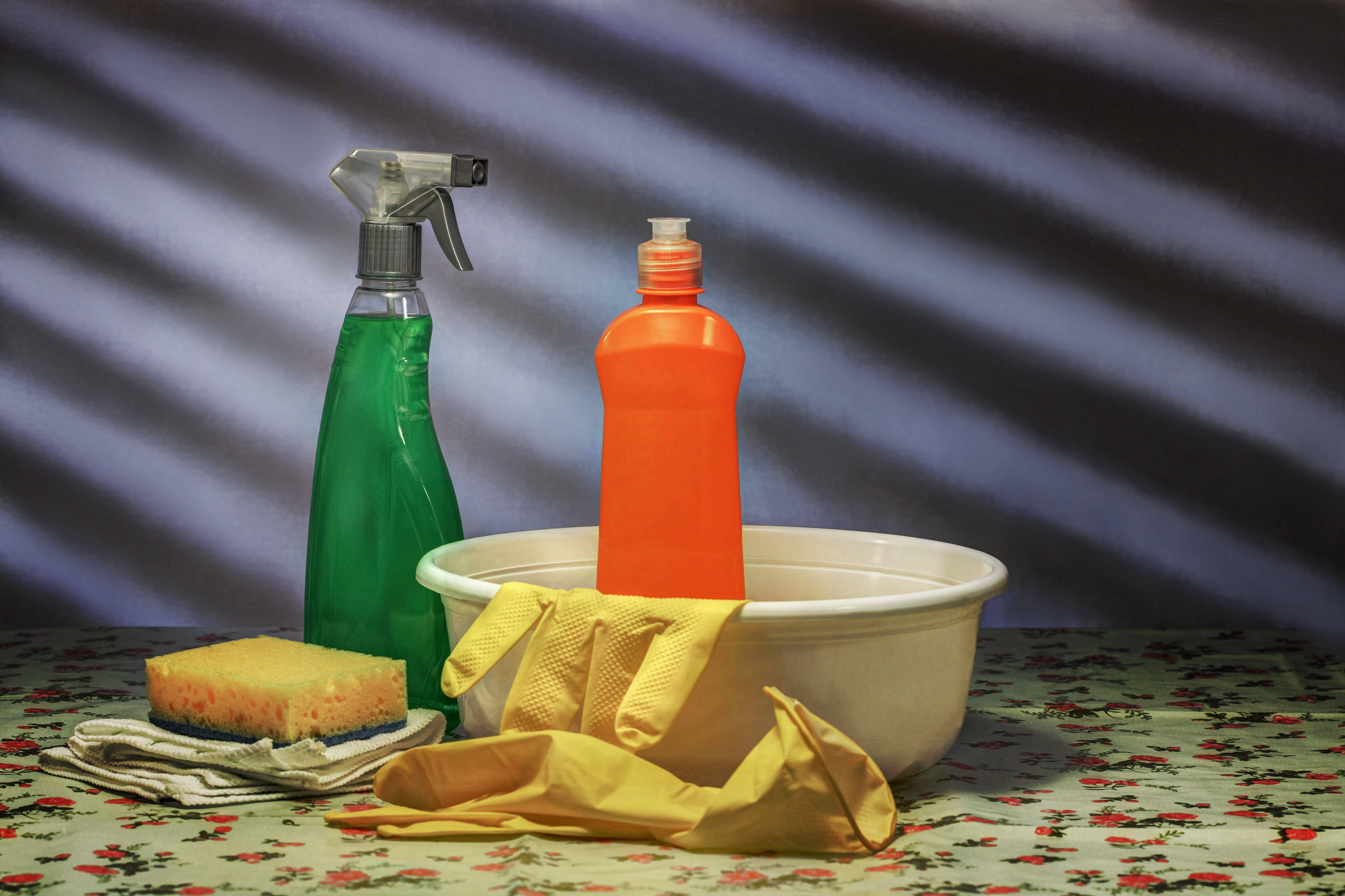 Cleaning 桌布 壁紙 — 一幅靜物畫，展示清潔用品，包括綠色噴霧瓶、橙色瓶子、黃色手套和海綿，排列在花卉圖案桌布上，背景為條紋。