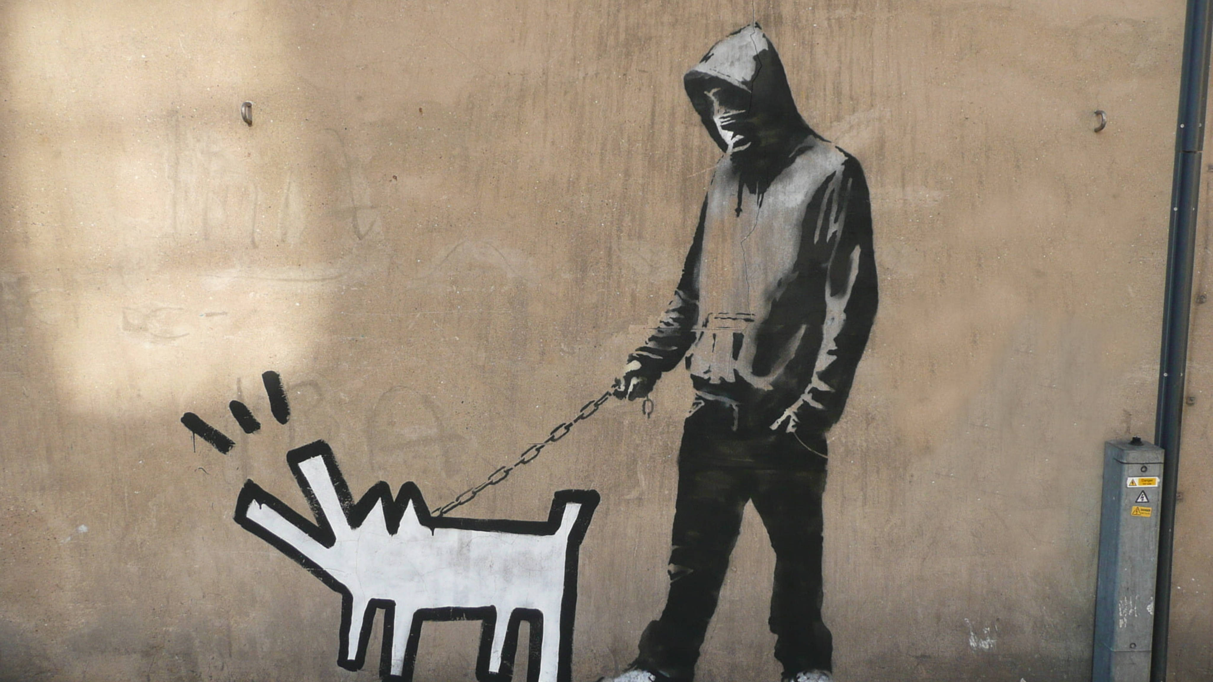Banksy 桌布 壁紙 — 一幅鮮明的黑白街頭壁畫描繪了一個戴兜帽的人物在風化的米色牆壁前用鏈條遛著一隻風格化的狗。