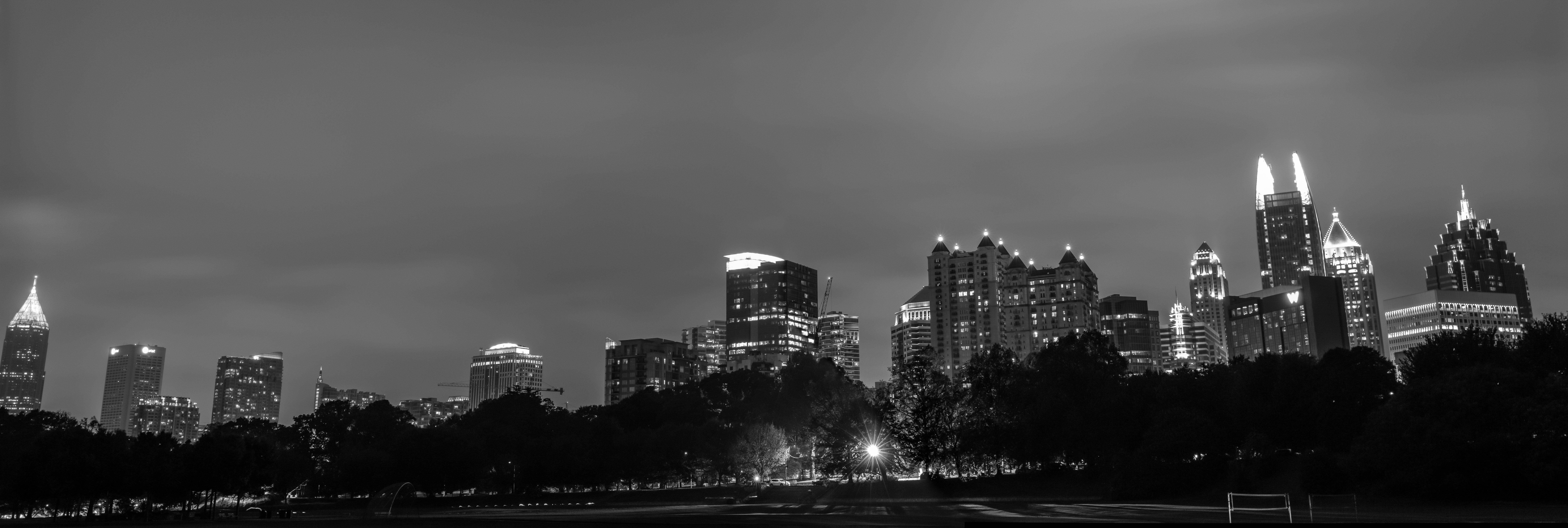 城市 桌布 壁紙 — 一幅黑白全景圖，展示夜晚的城市天際線，前景有燈火通明的摩天大樓和黑暗的樹木剪影。
