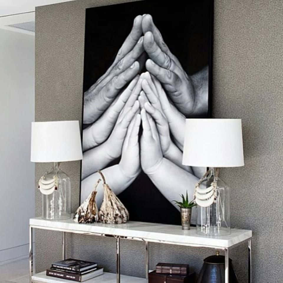 Praying 桌布 壁紙 — 一幅黑白攝影藝術印刷品，描繪層疊的祈禱之手，懸掛在現代玄關桌上，兩旁各有一盞玻璃燈。
