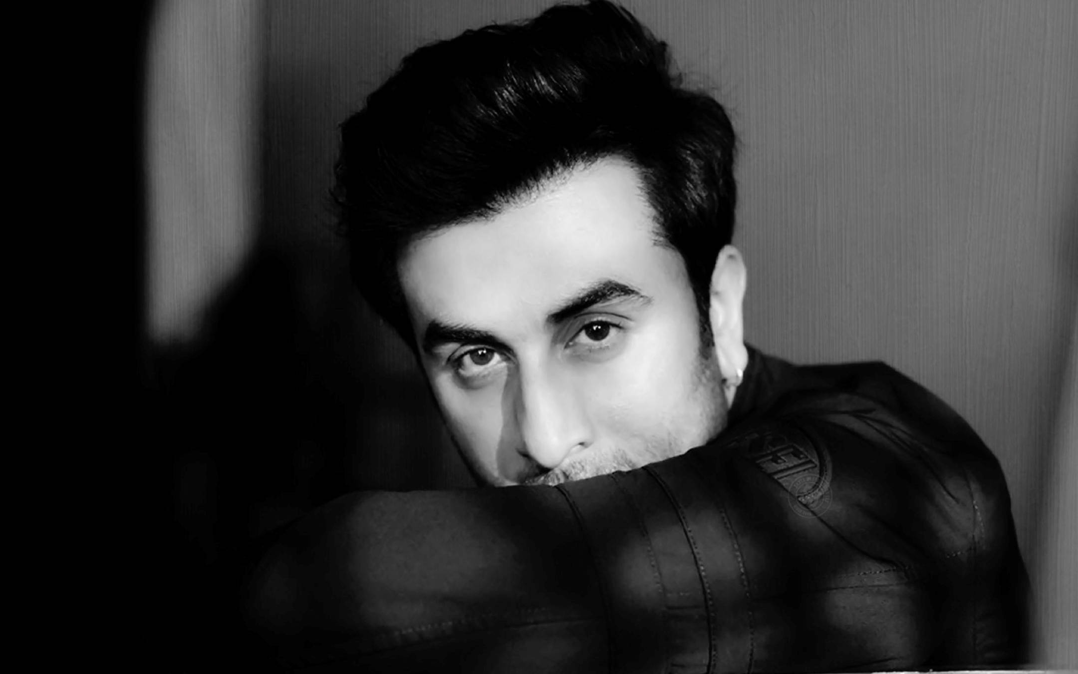 Ranbir Kapoor 桌布 壁紙 — 一幅黑白肖像，一名穿著皮夾克的男子向前傾，眼神銳利，背景是紋理牆壁。