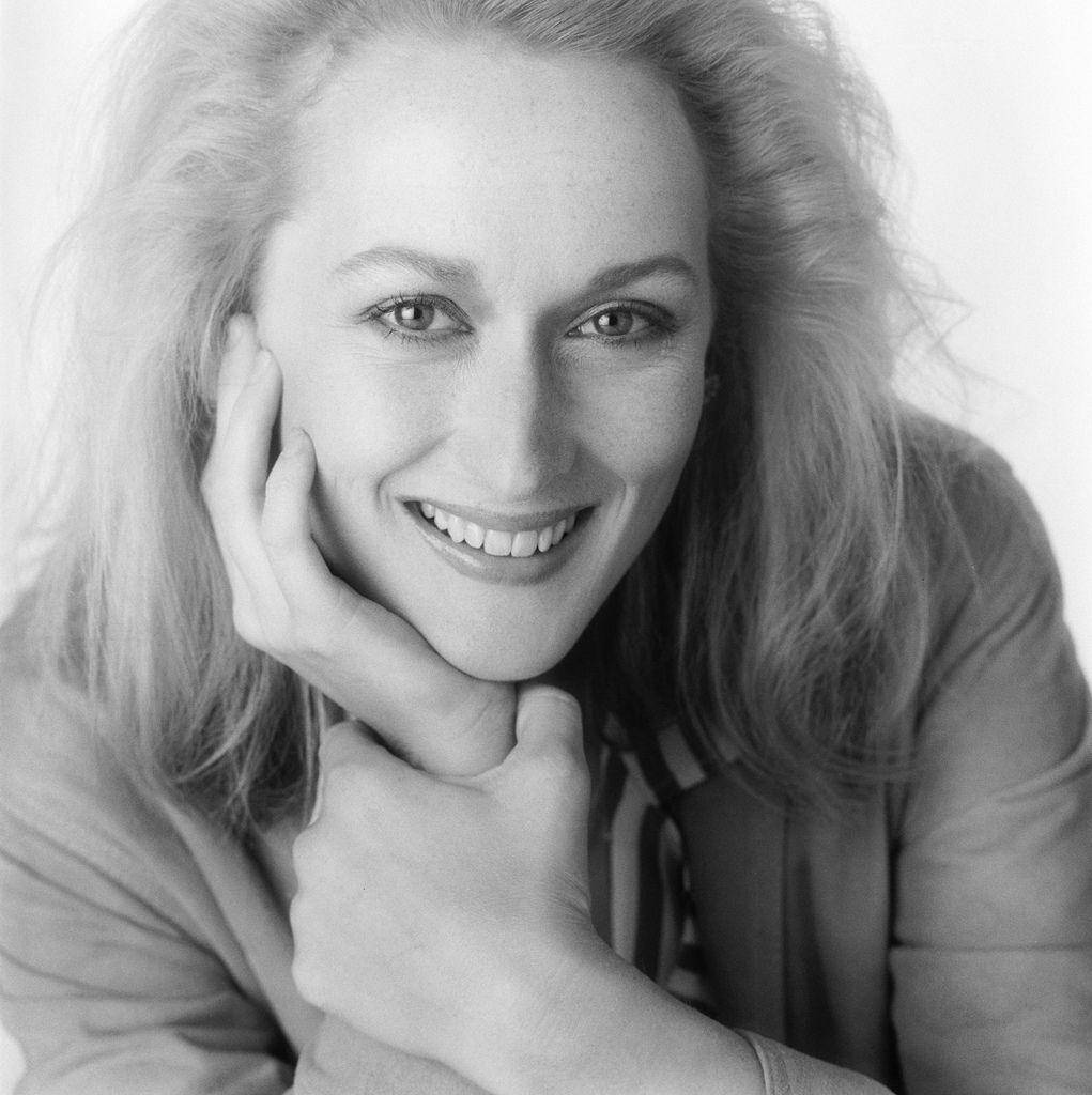Meryl Streep 桌布 壁紙 — 一幅黑白肖像照，一位微笑的波浪髮女性在純色背景前將下巴擱在手上。