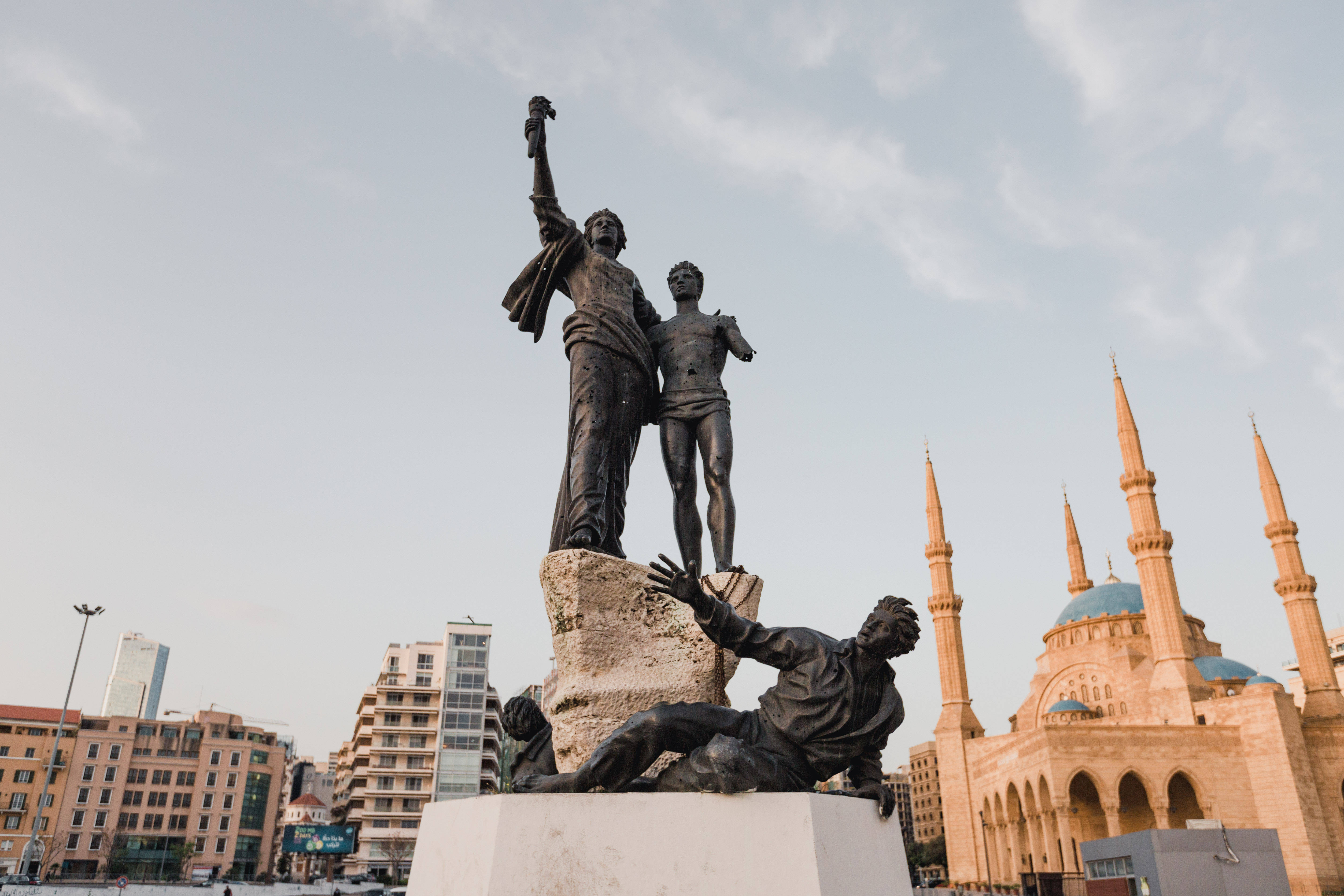 Lebanon 桌布 壁紙 — 一座三位人物的銅像，一人高舉手臂站立，另一人跪在倒地的人物旁，背景是宏偉的清真寺和現代建築，天空柔和。