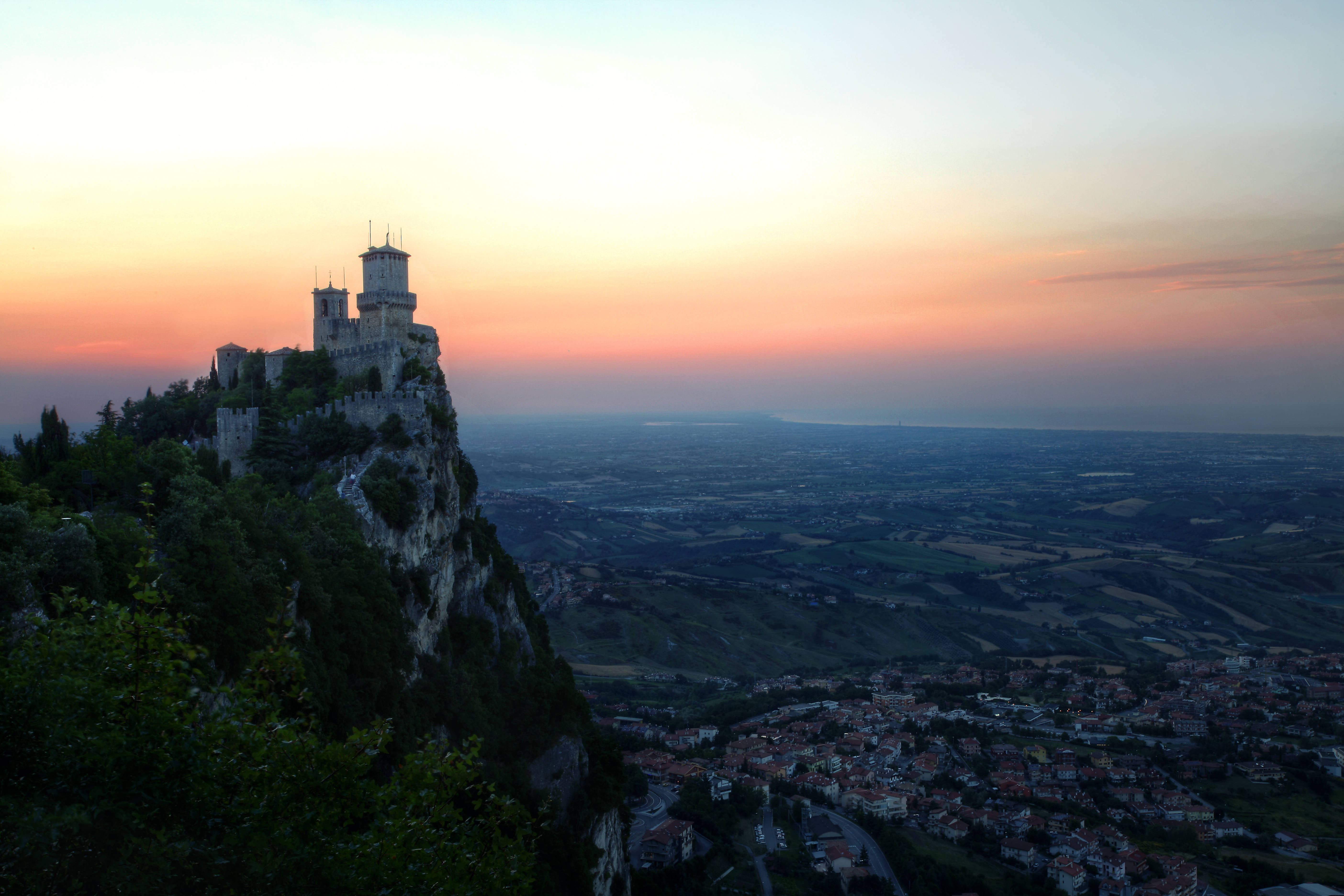 San Marino 桌布 壁紙 — 一座中世紀城堡坐落在日落時分的懸崖頂上，俯瞰著下方廣闊的山谷和小鎮。