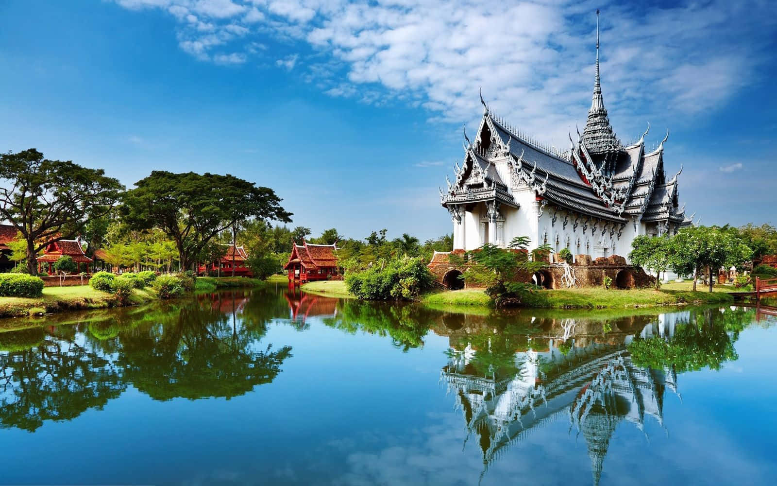 View 桌布 壁紙 — 一座令人驚嘆的泰國寺廟，擁有精緻的尖頂，在明亮的天空下完美地倒映在平靜的藍色池塘中。