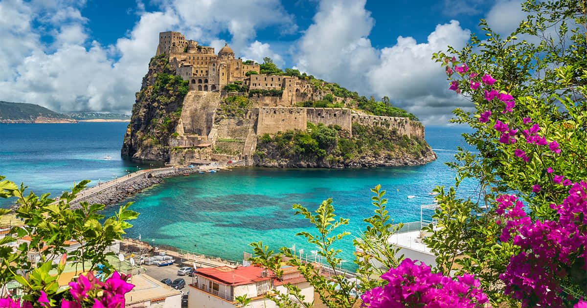 Italian Island 桌布 壁紙 — 一座令人驚嘆的海岸堡壘，坐落在岩石島上，周圍環繞著碧綠的海水，並以鮮豔的粉紅色花朵作為框架。